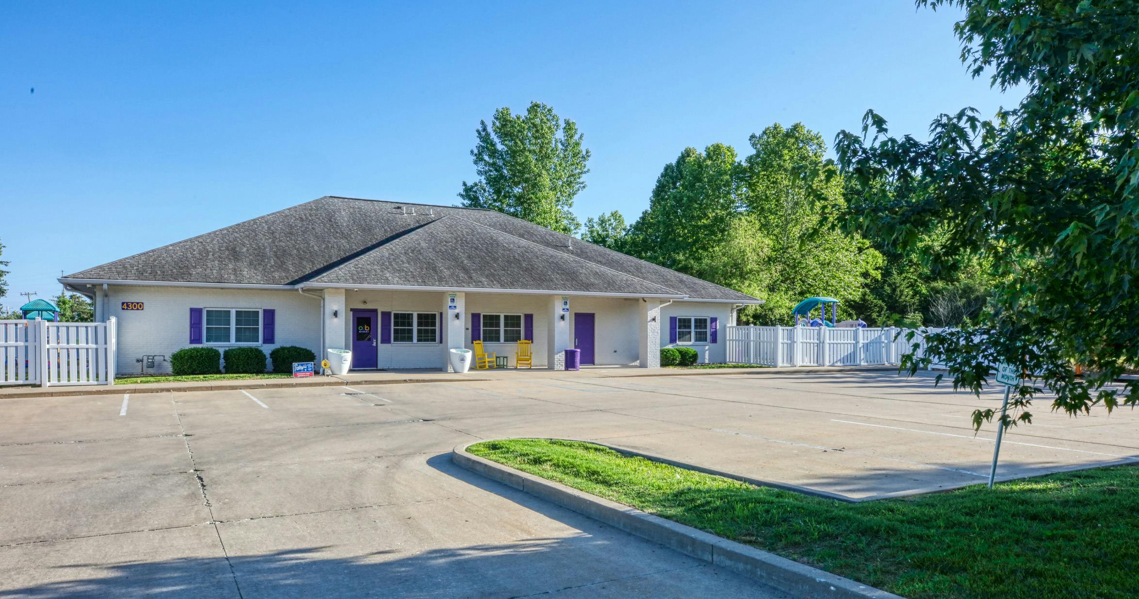 White building with purple doors and shutters, a parking lot, and a fenced playground