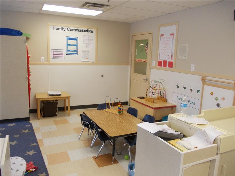 An empty classroom with small tables and chairs, a bulletin board, and toys