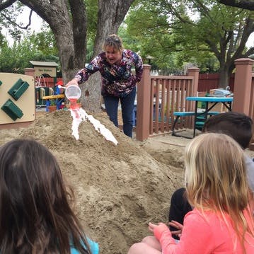 Adult pouring liquid into a sand volcano with children watching