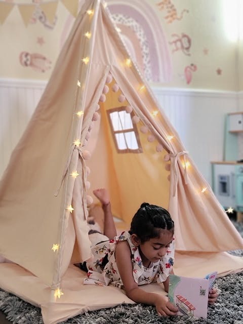 Child lying inside a teepee tent with string lights and a book