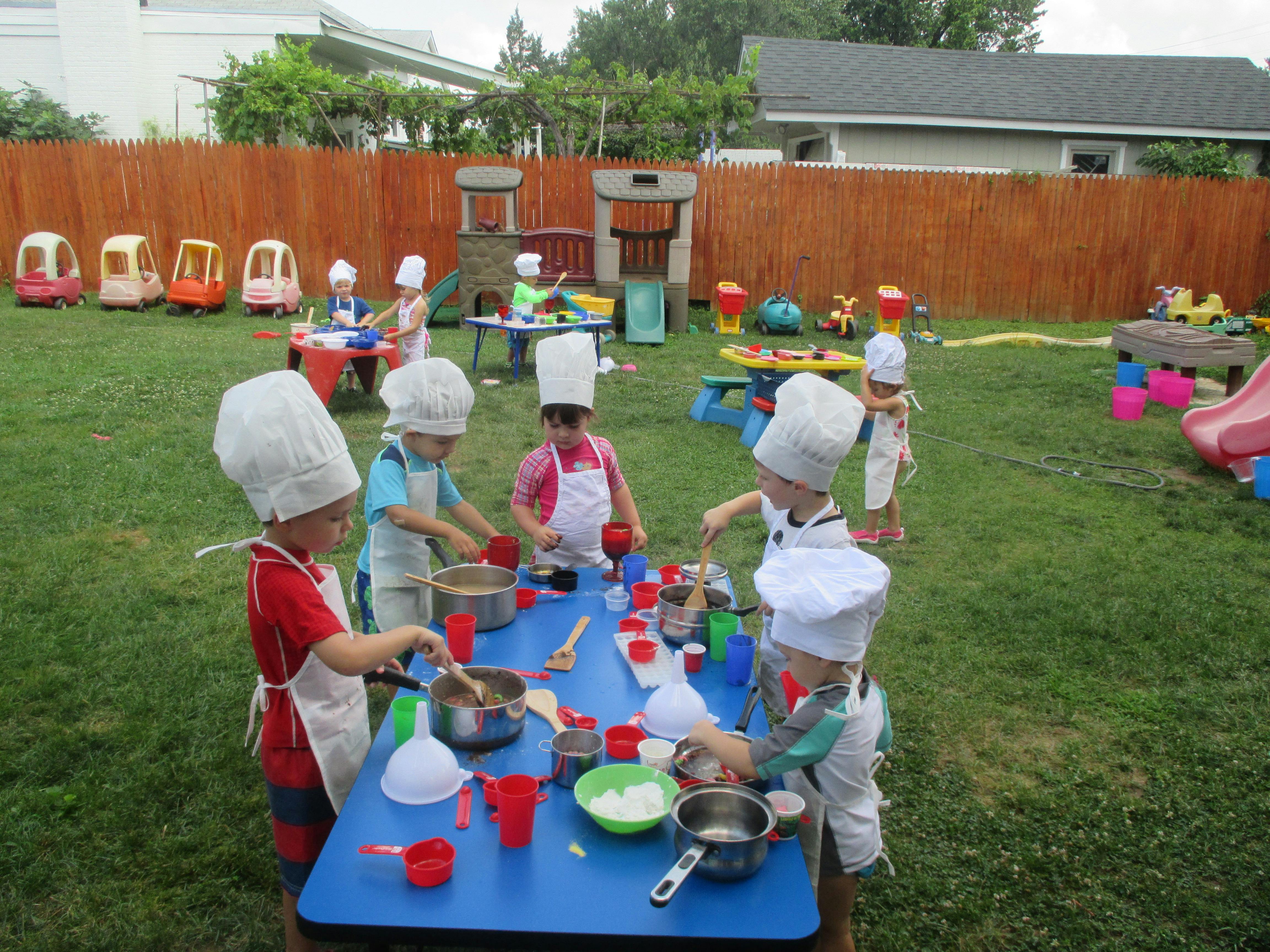 Children in chef hats and aprons mixing ingredients at a table outdoors