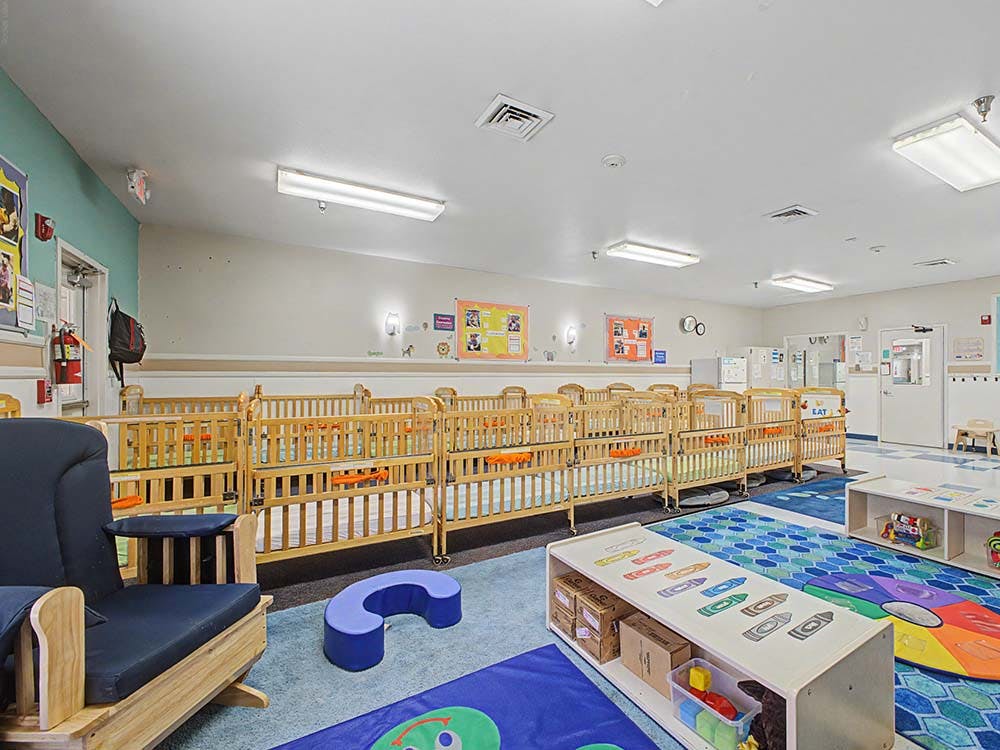 A room with multiple wooden cribs lined up, a rocking chair, and play mats