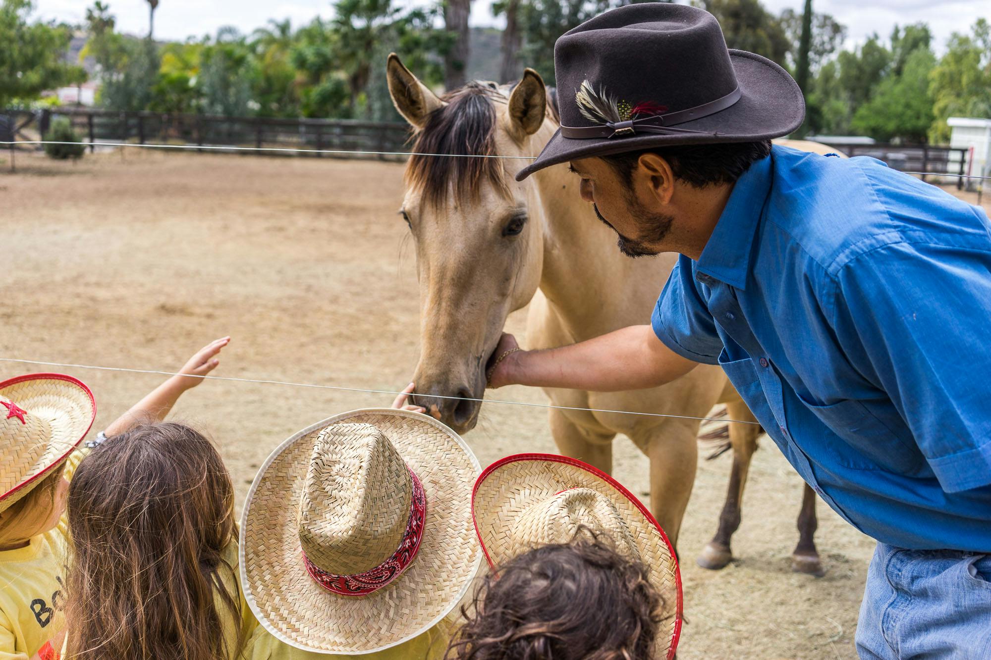 Person in a hat petting a horse while children in hats watch