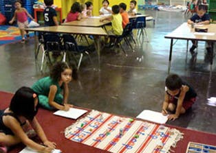 Children writing on paper on the floor and at tables in a classroom