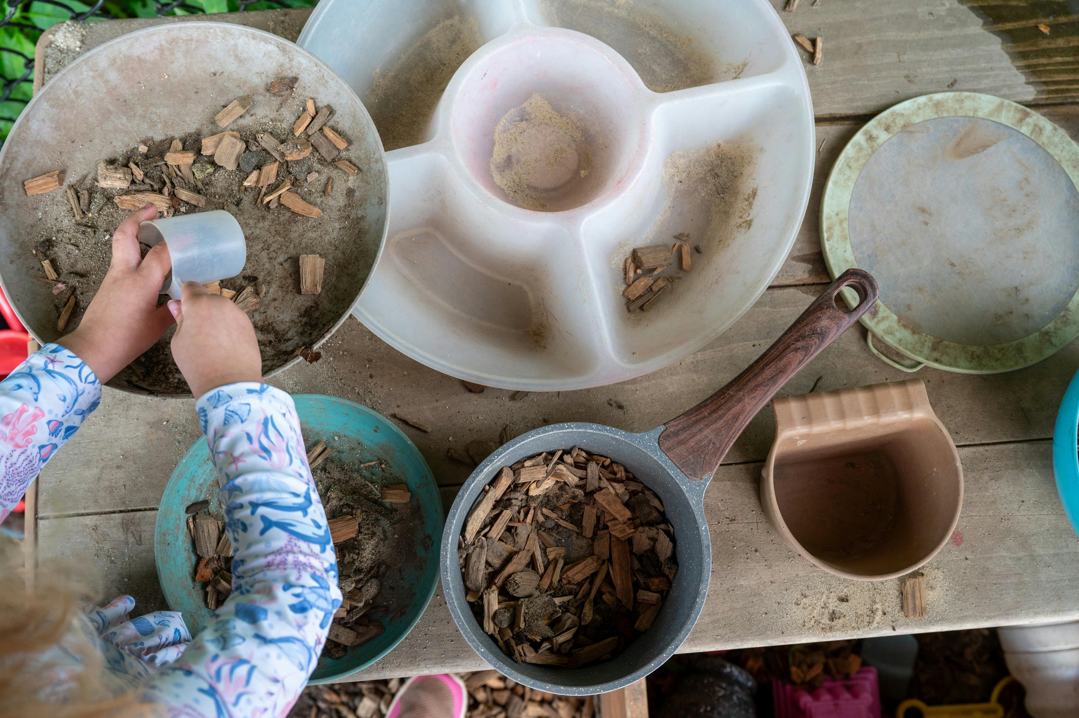 Child playing with wood chips and sand in trays on a wooden table
