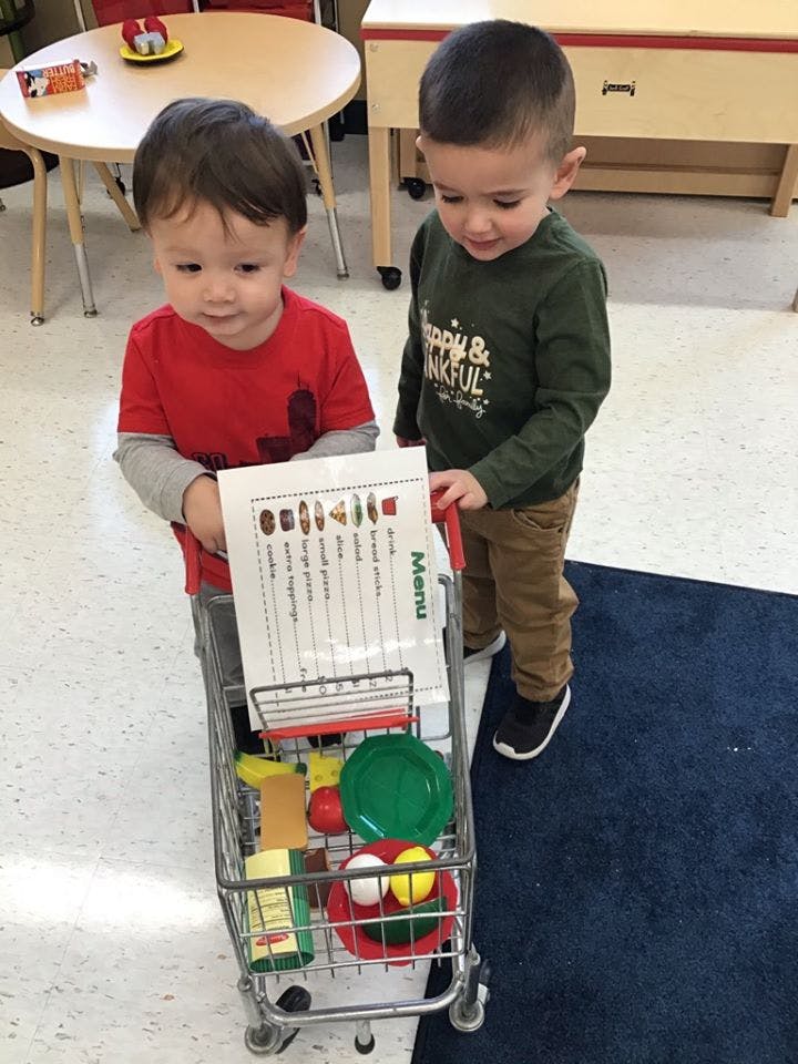 Two children pushing a toy shopping cart with a menu and play food