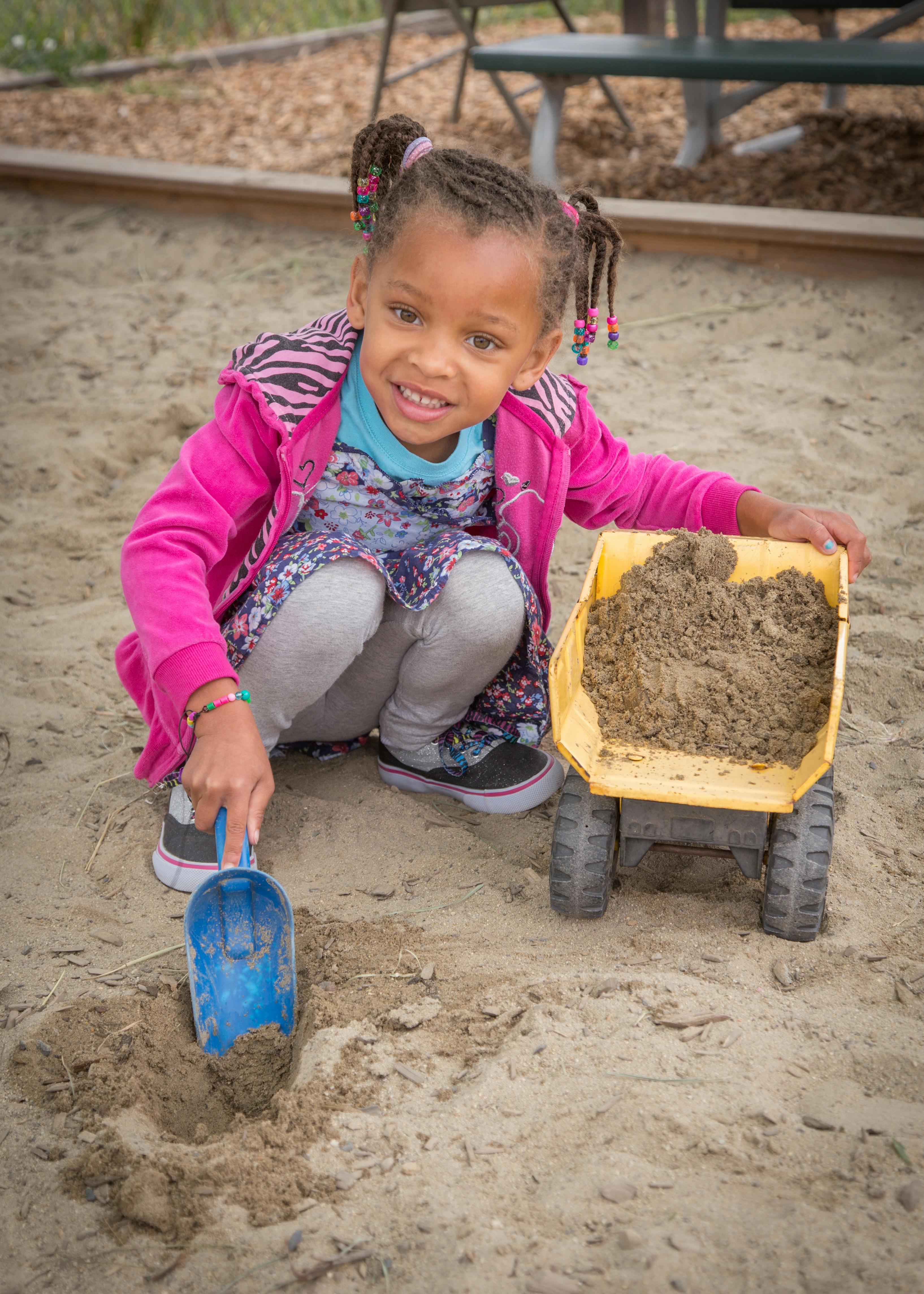 Child playing in sand with a blue shovel and yellow toy truck