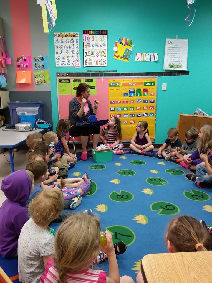 Adult speaking to a group of children sitting on a rug in a classroom