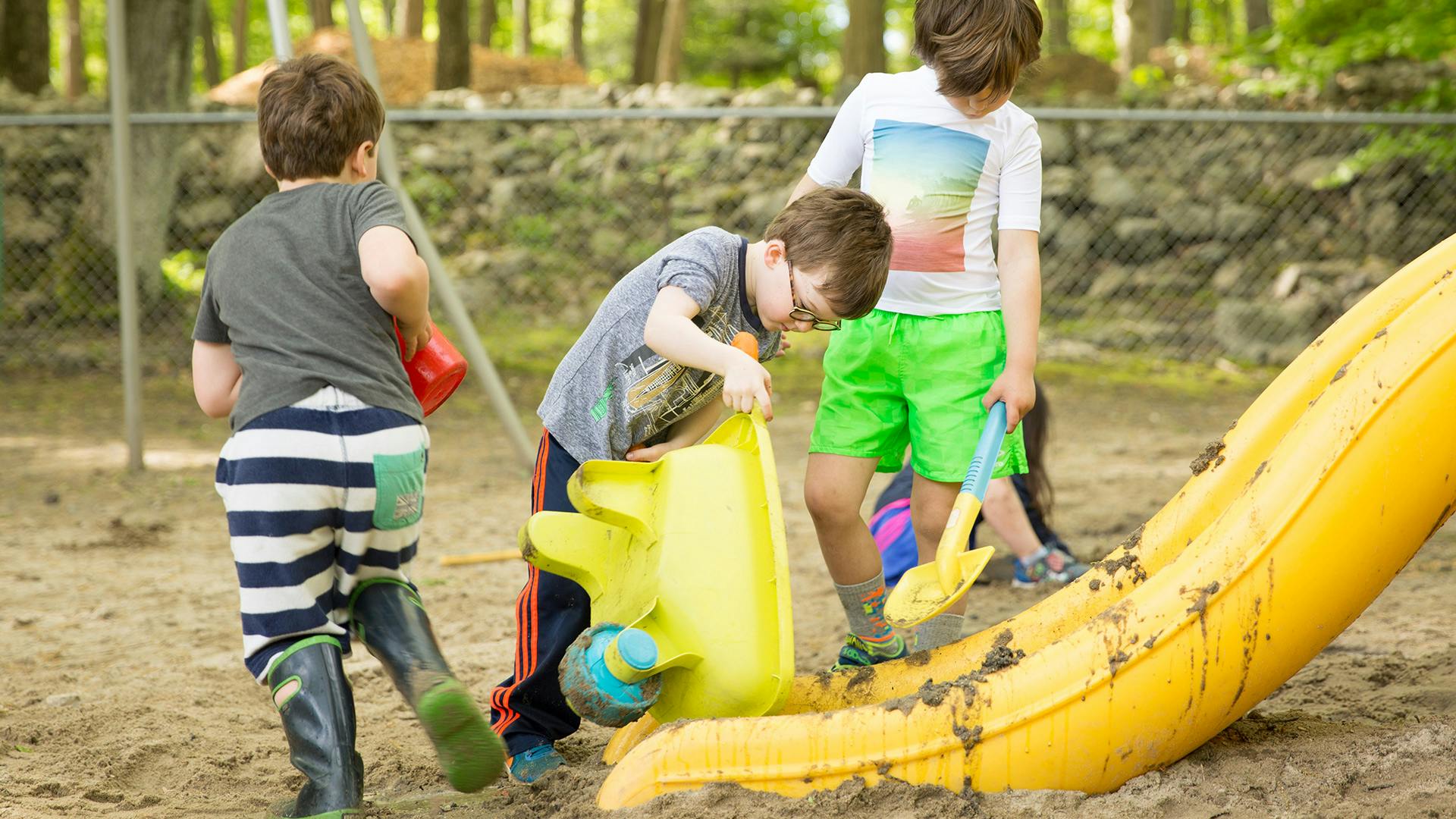 Three children playing in sand with toys and a yellow slide