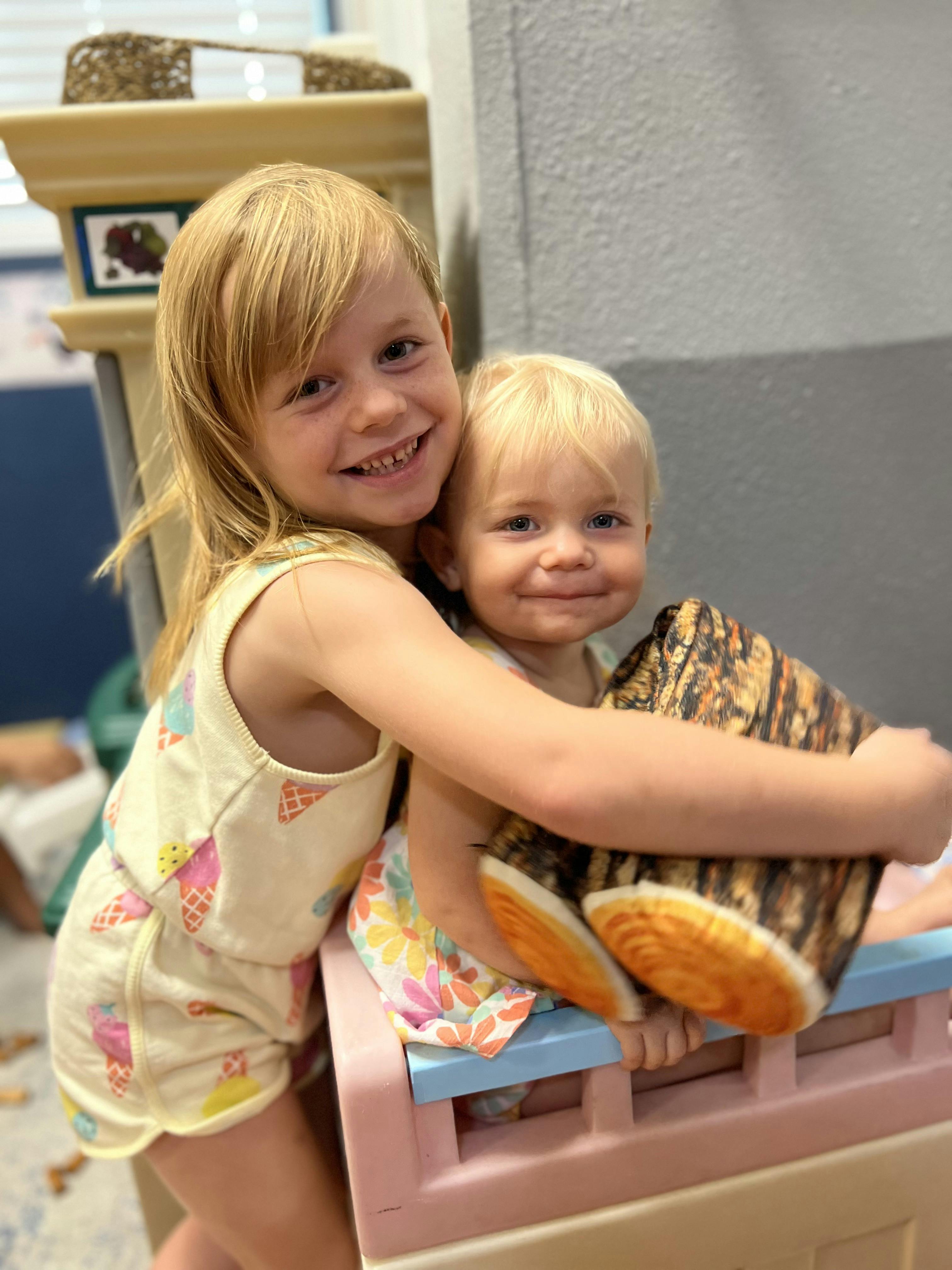 Child hugging a younger child holding a log-shaped pillow