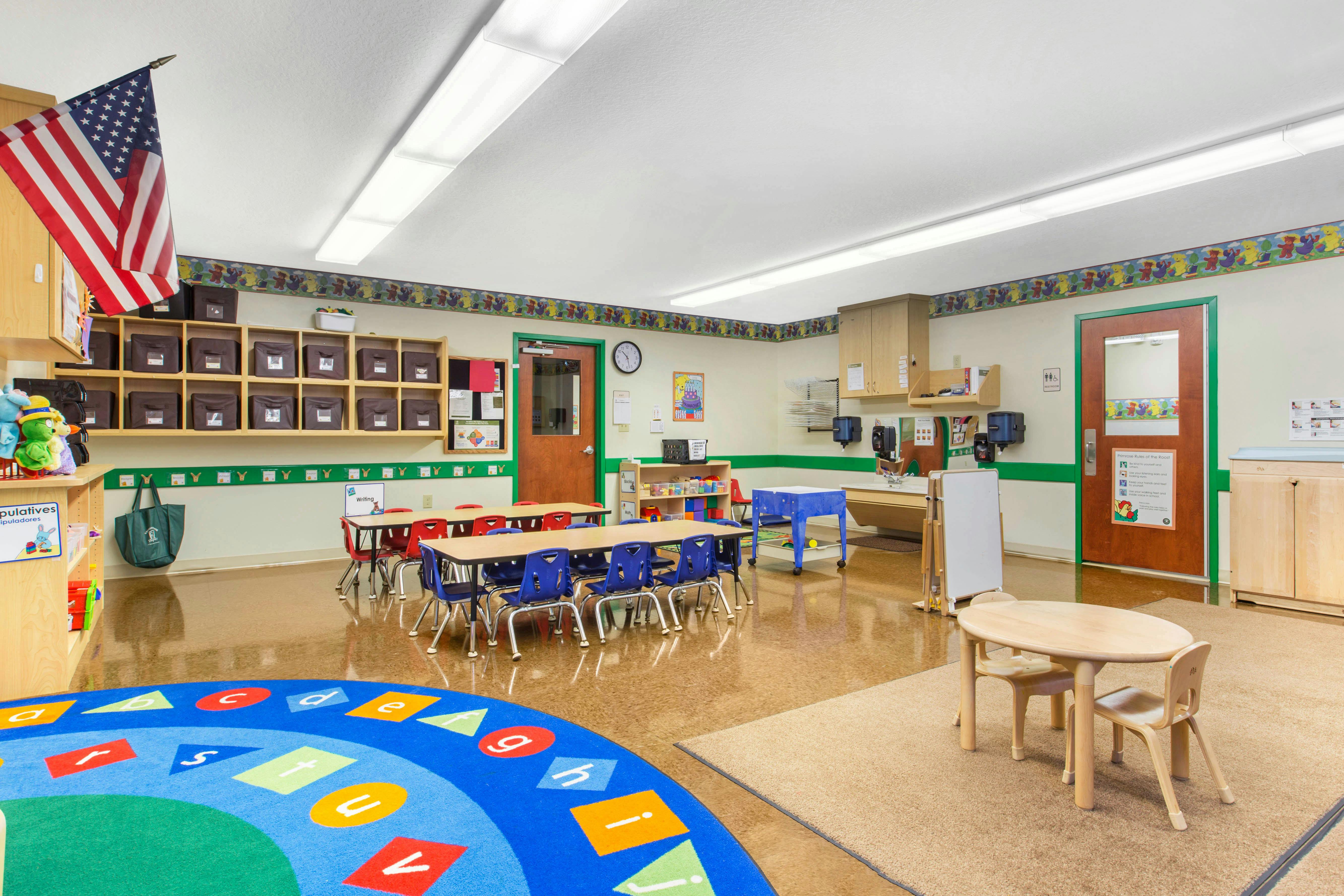 Empty classroom with small tables and chairs, a colorful rug, and an American flag