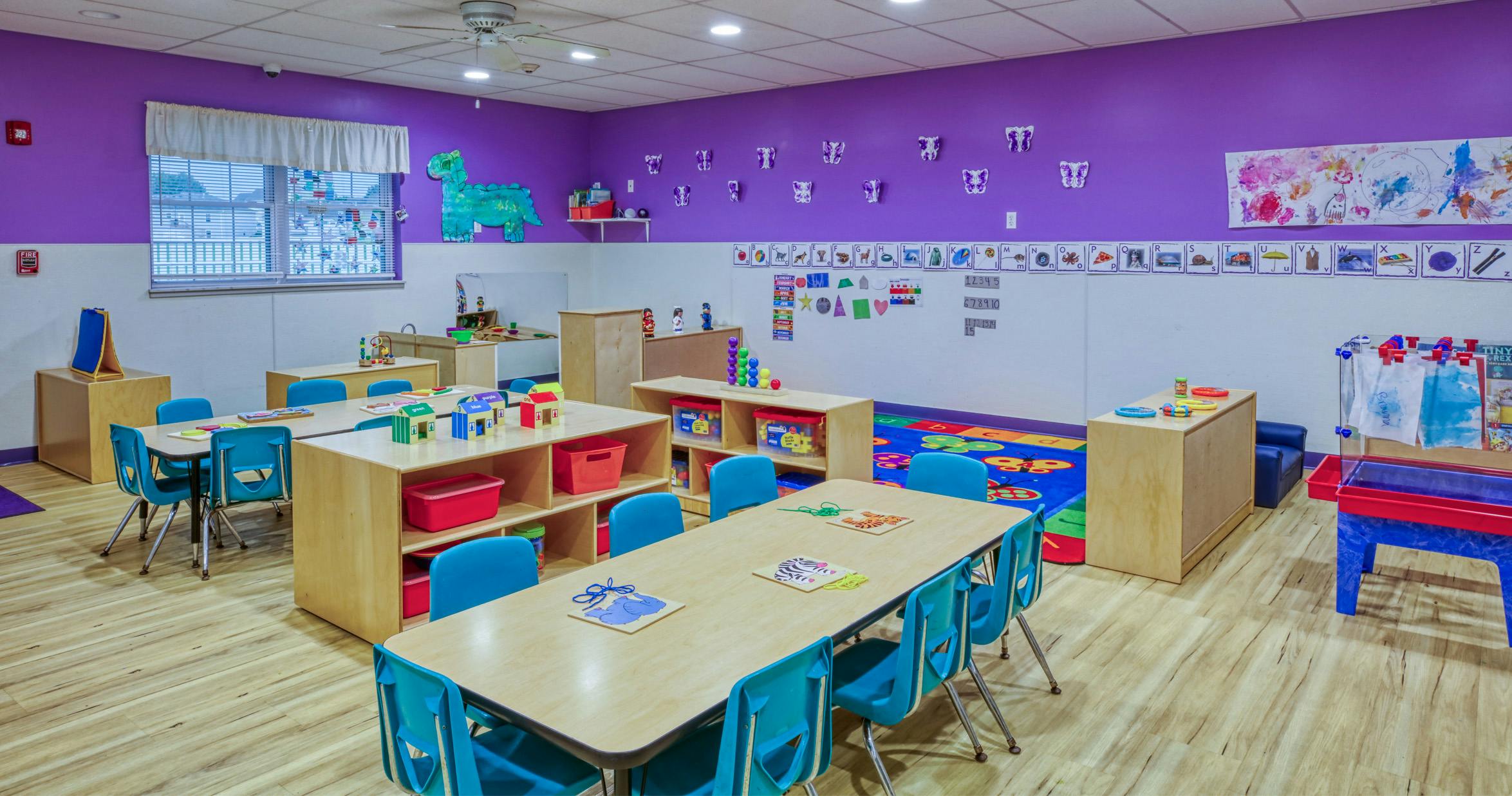An empty children's classroom with purple walls, small tables, and blue chairs