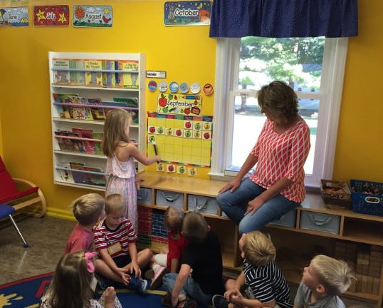 Adult and children in a classroom, child pointing at a calendar