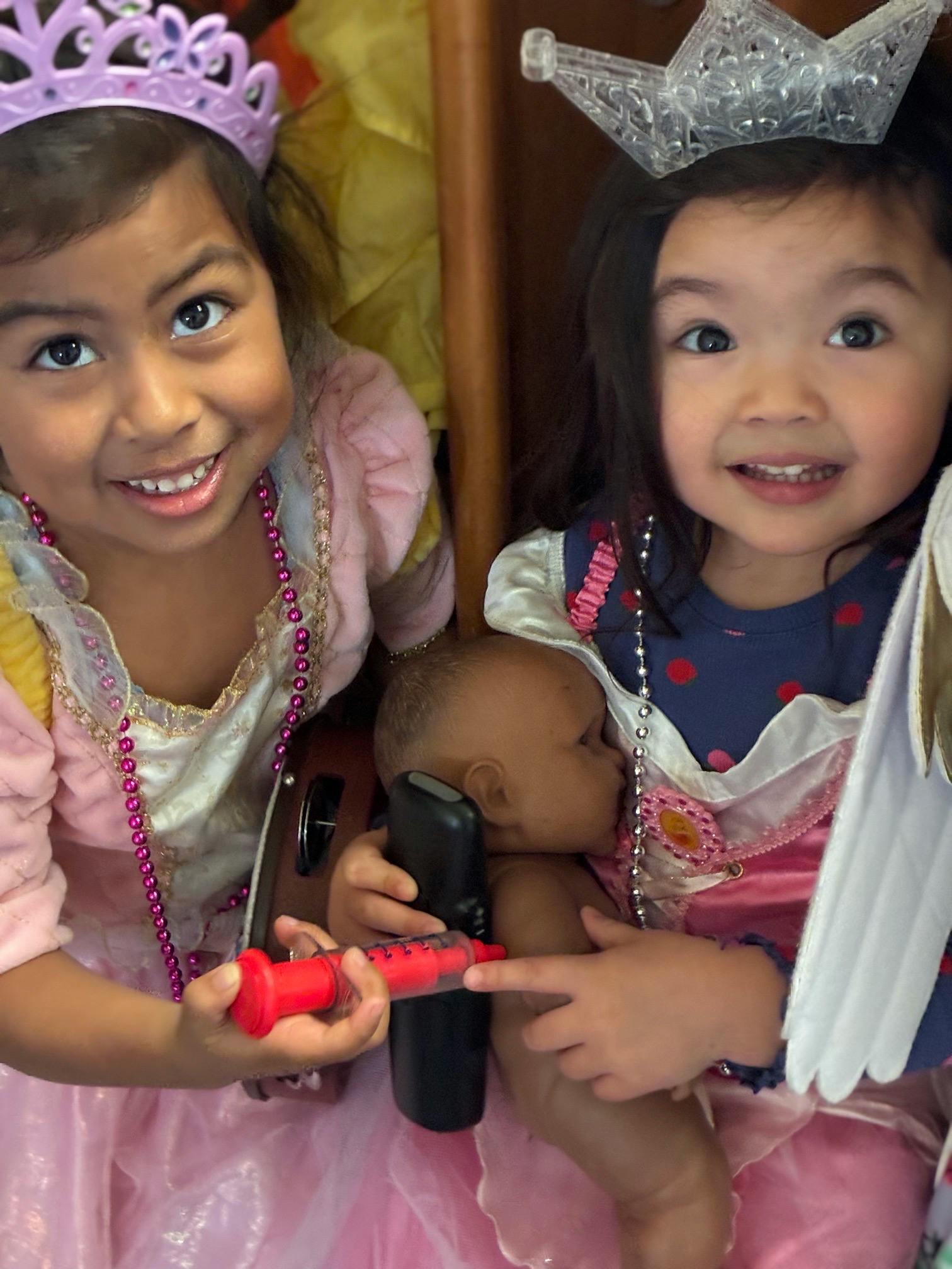 Two children wearing crowns playing with a doll and a toy syringe