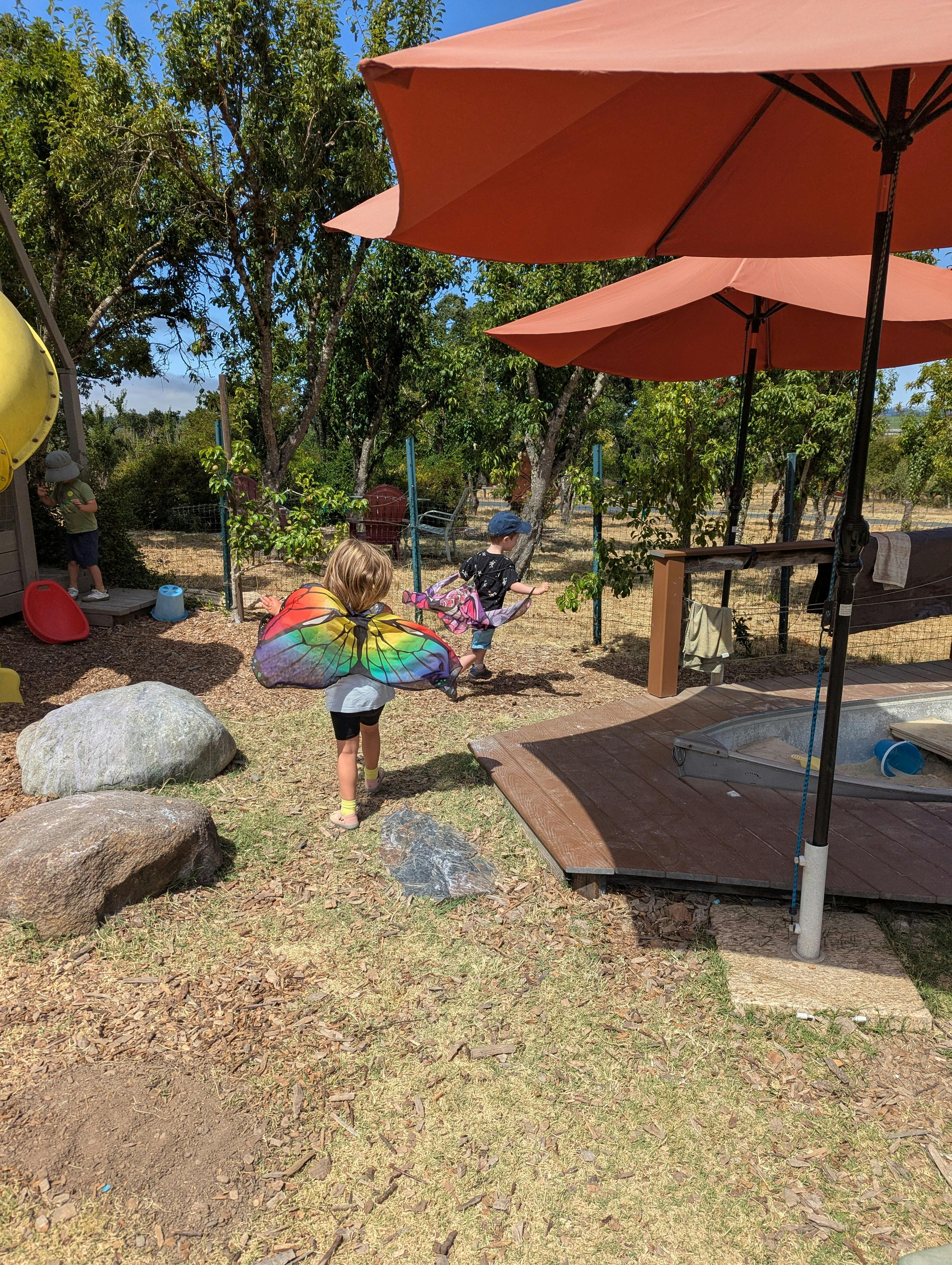 Child with rainbow wings walking on grass near umbrellas