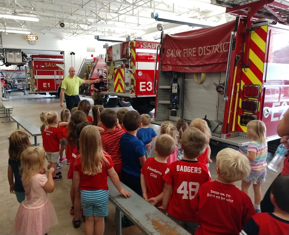 Adult speaking to a group of children in front of fire trucks