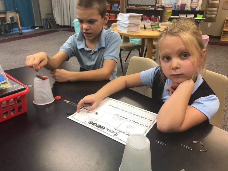 Two children at a table, one working with a cup, the other looking forward