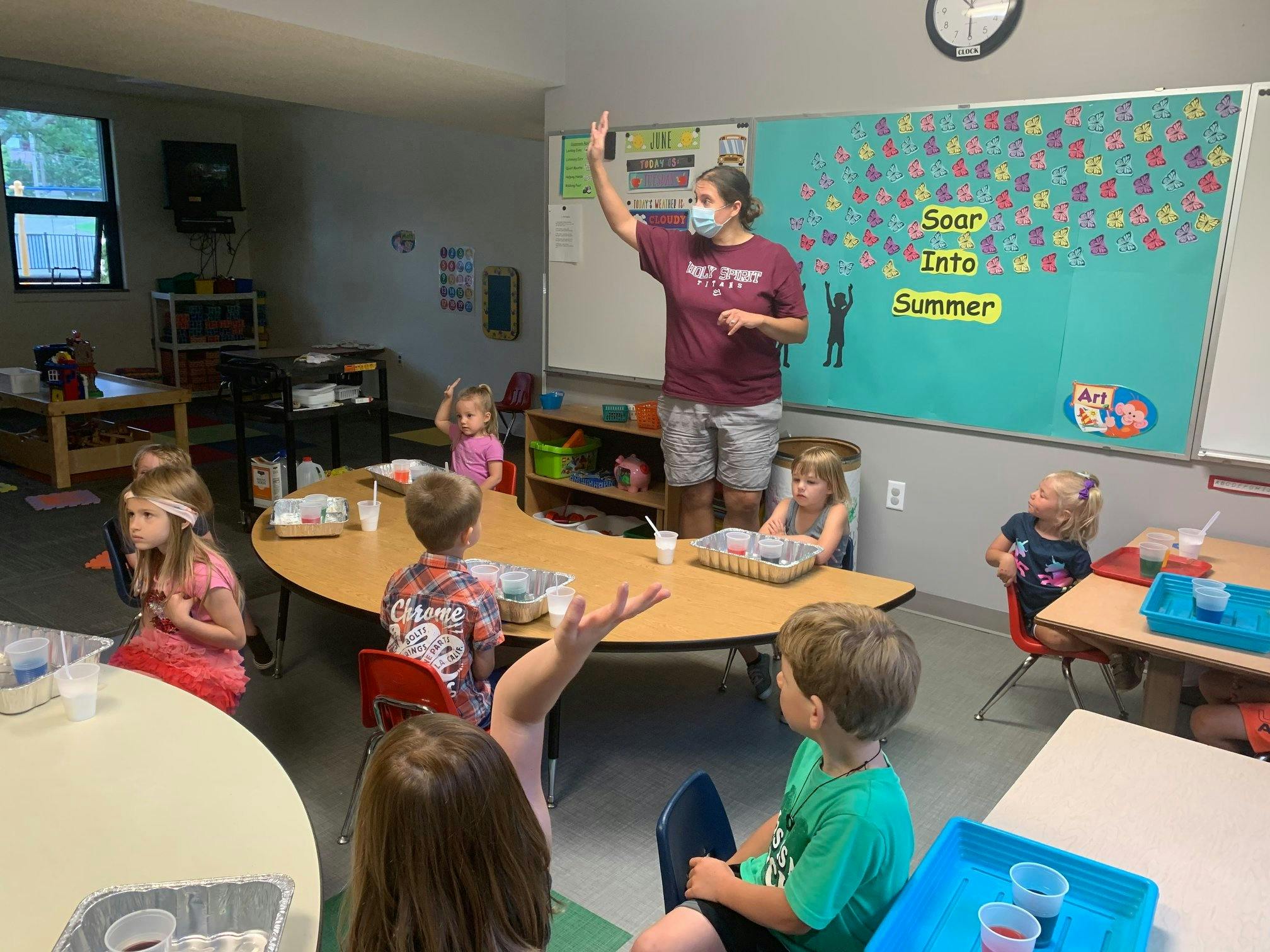 Person in mask raising hand in a classroom with children seated children