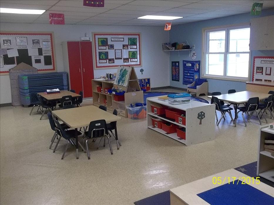 An empty classroom with small tables and chairs, shelves with red bins, and bulletin boards