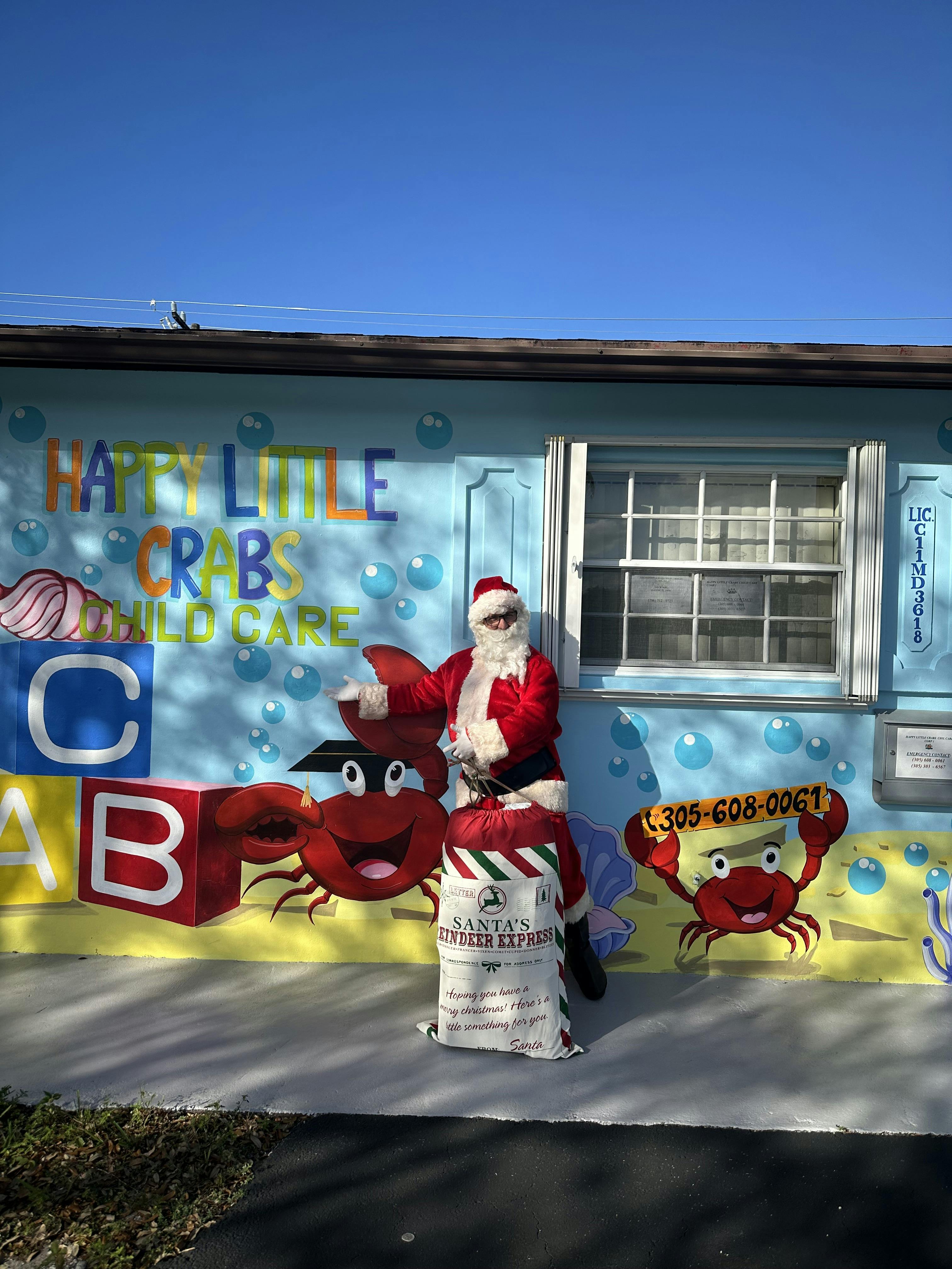 Person dressed as Santa Claus standing next to a bag in front of a colorful mural