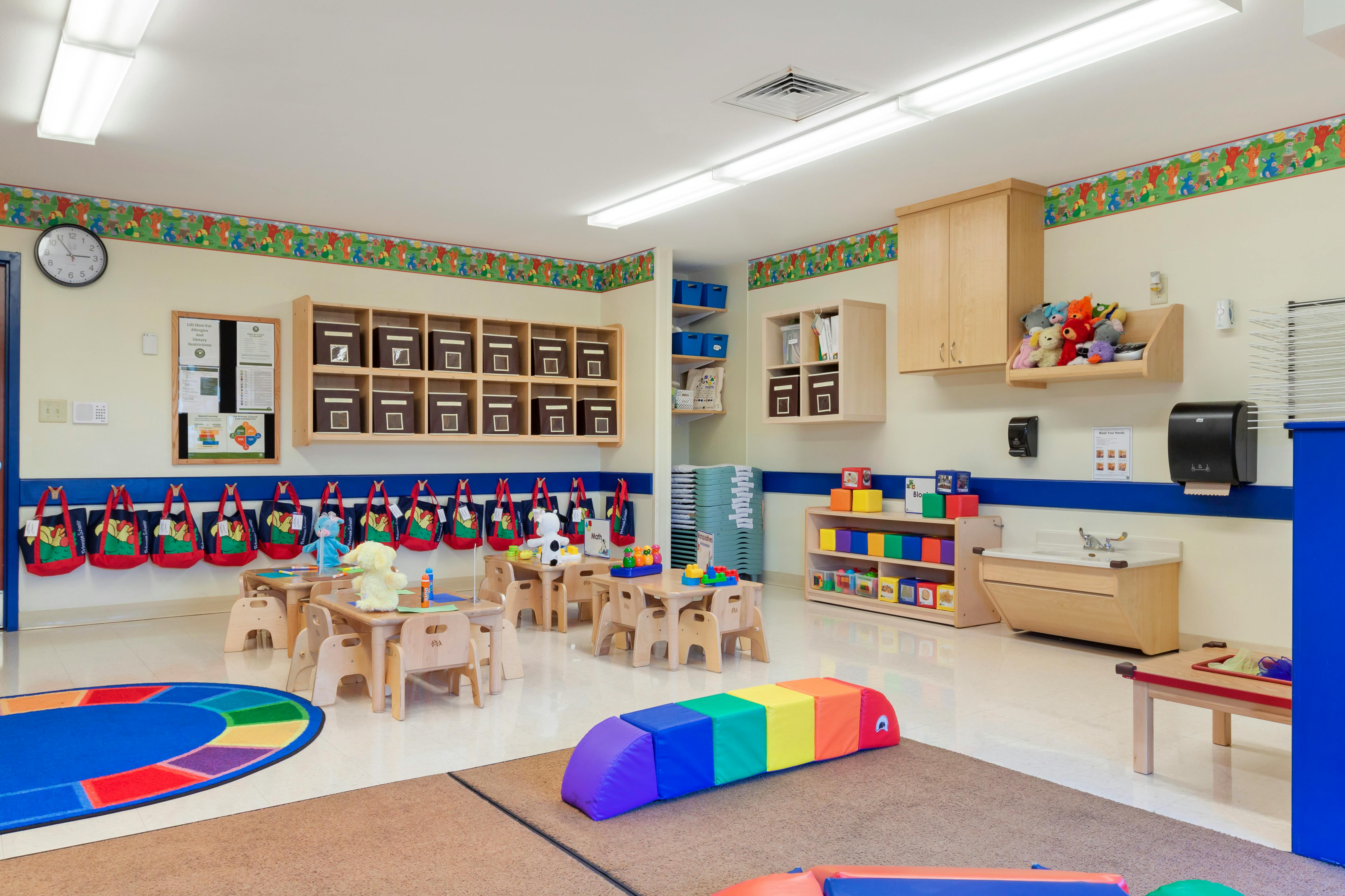 Classroom with small tables and chairs, shelves with storage boxes, and a rainbow play mat