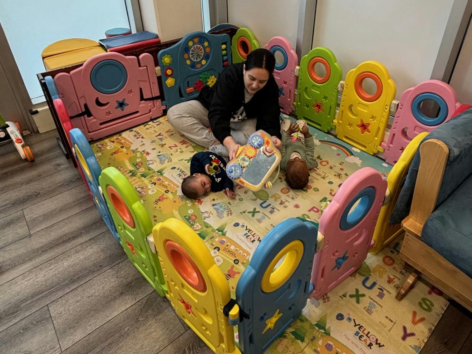 person playing with two babies on a colorful playmat inside a playpen