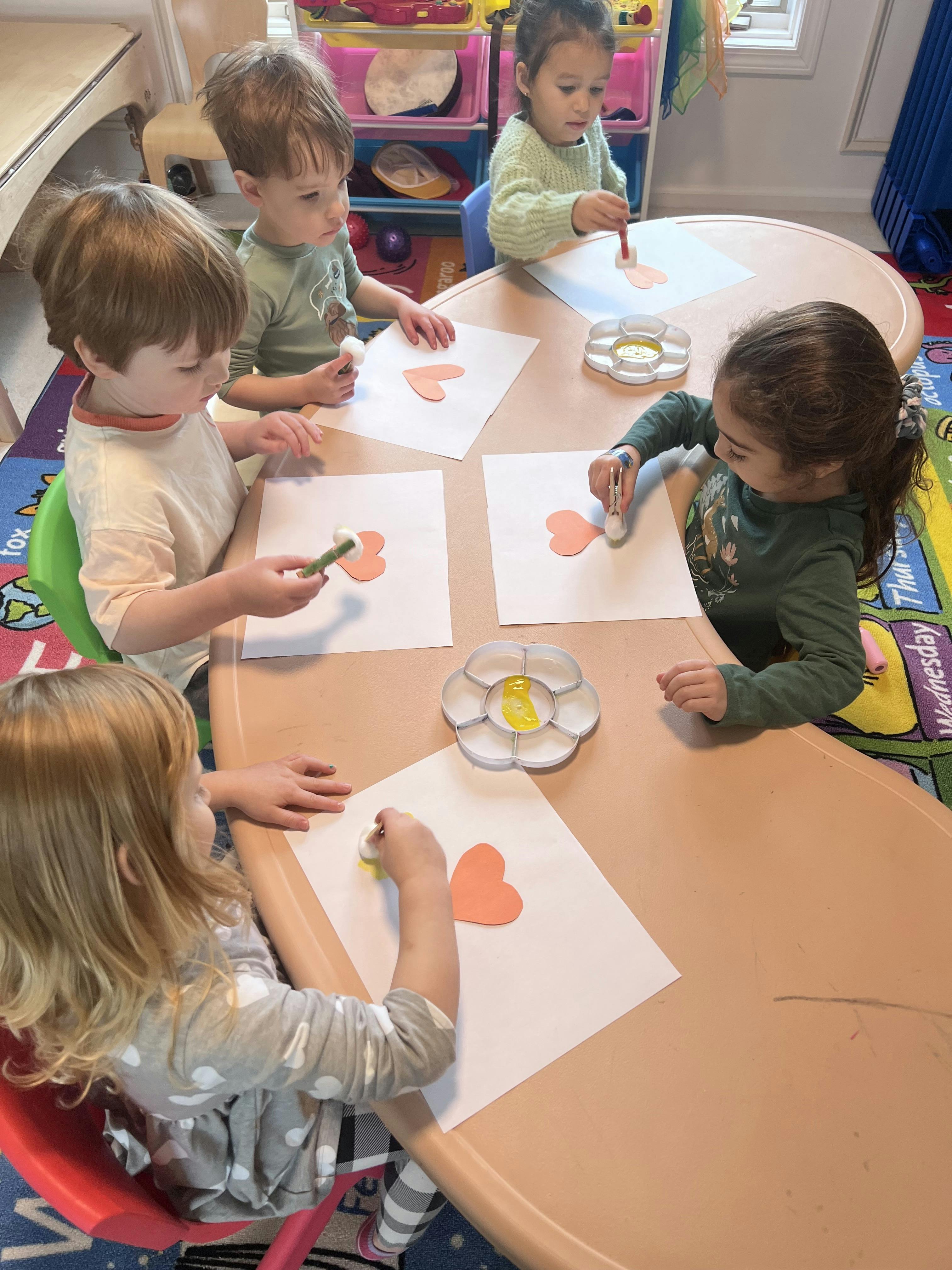 Four children painting hearts on paper with cotton swabs at a table