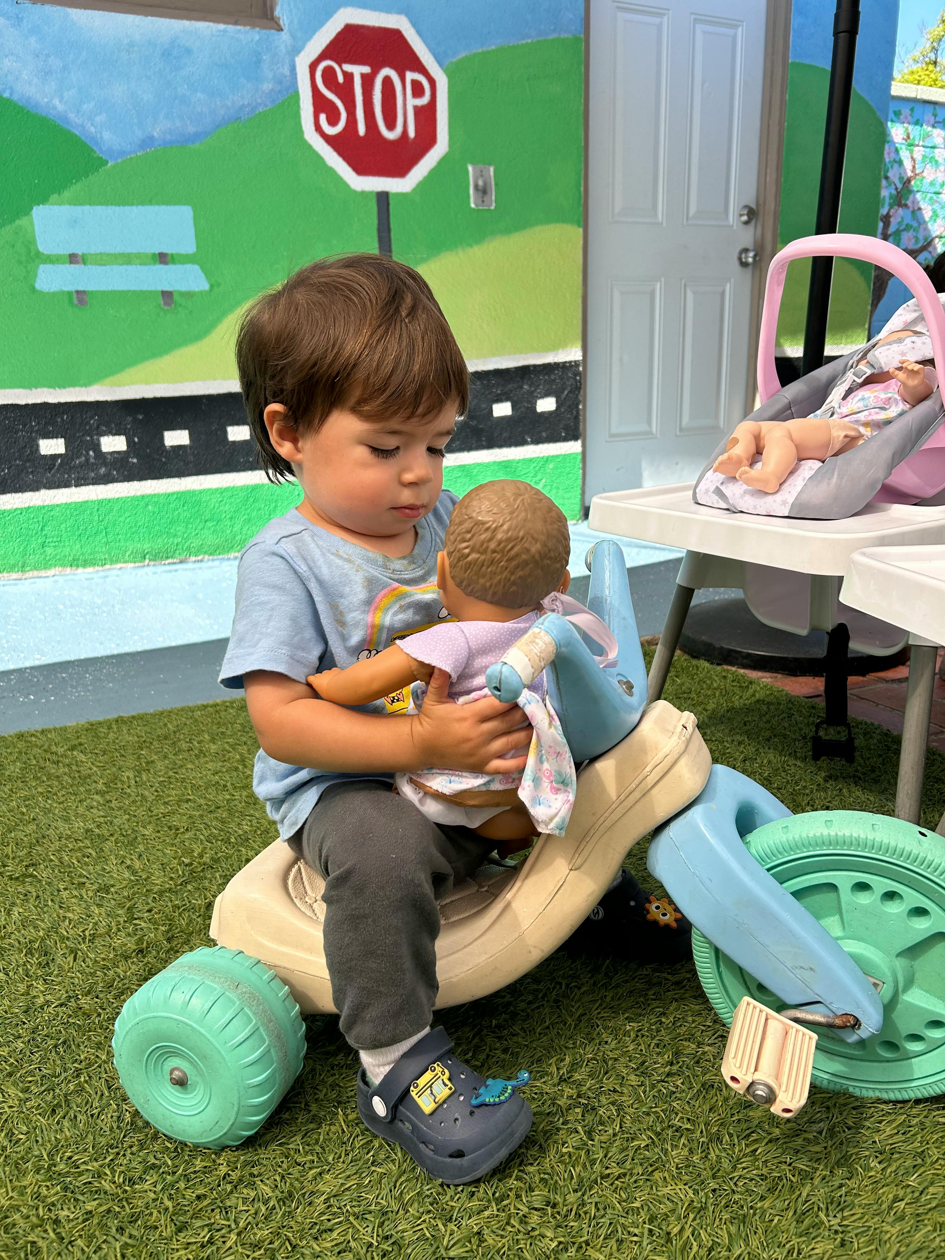 Child sitting on a toy tricycle holding a doll