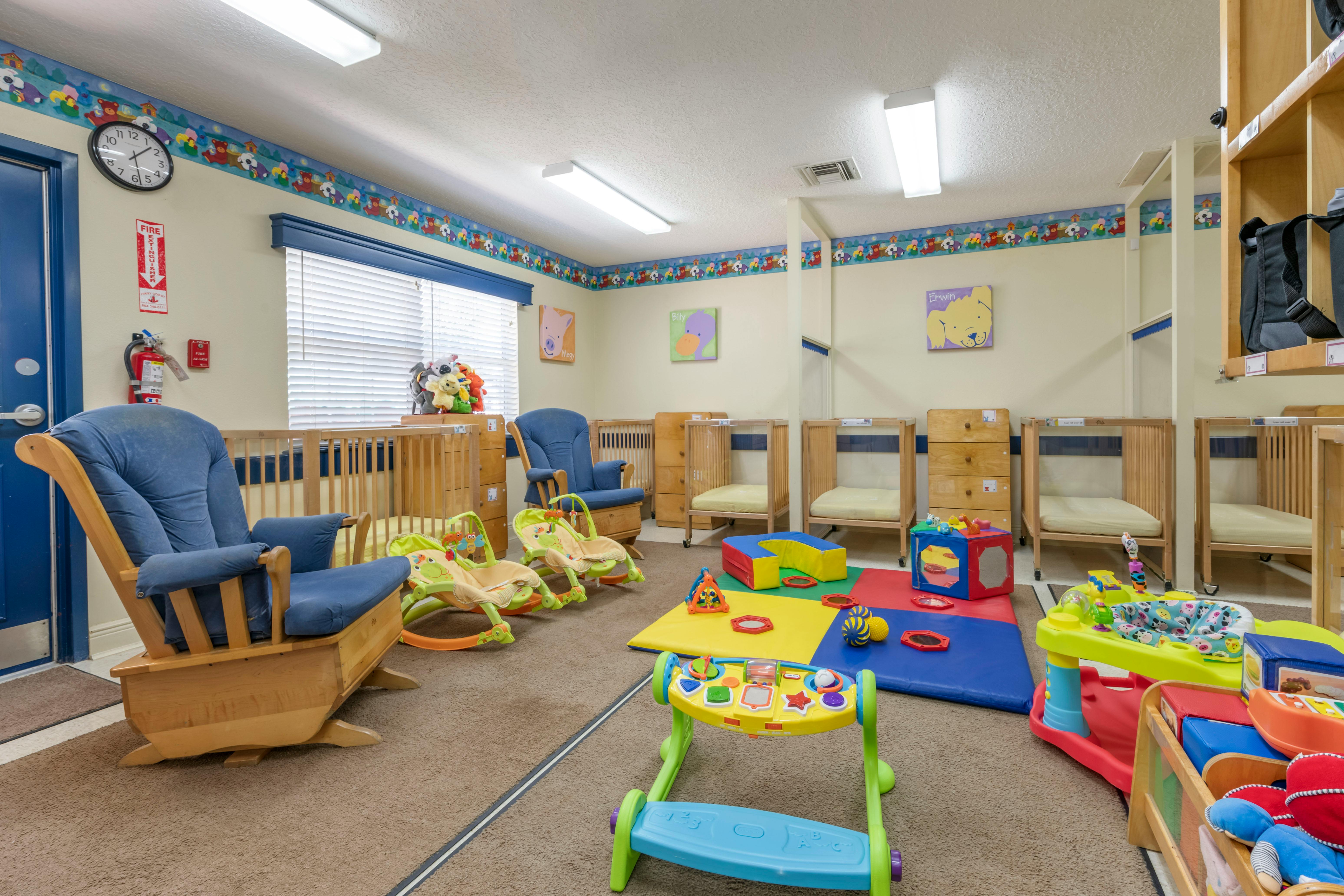 A colorful childcare room with cribs, rocking chairs, play mats, and baby toys