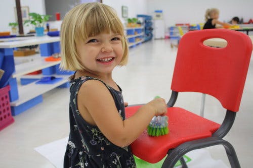 Child cleaning a red chair with a brush in a classroom