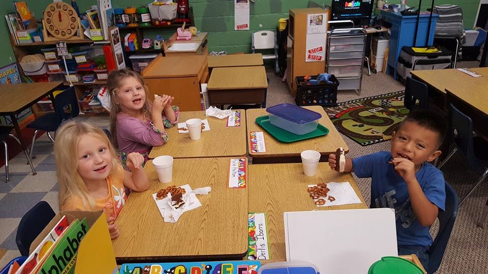 Three children sitting at desks in a classroom eating snacks