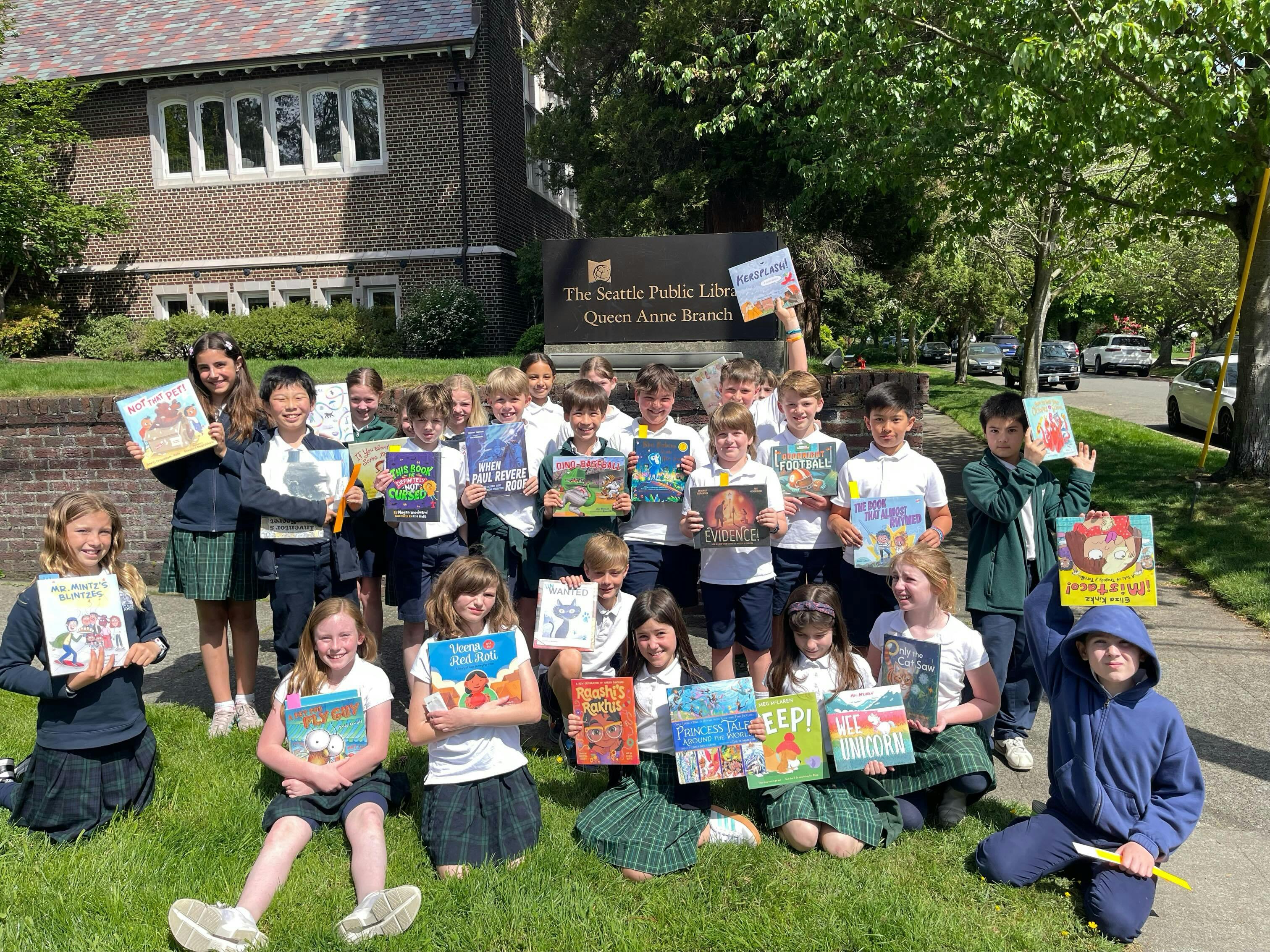 Group of children holding books in front of a library sign that reads "The Seattle Public Library Queen Anne Branch"