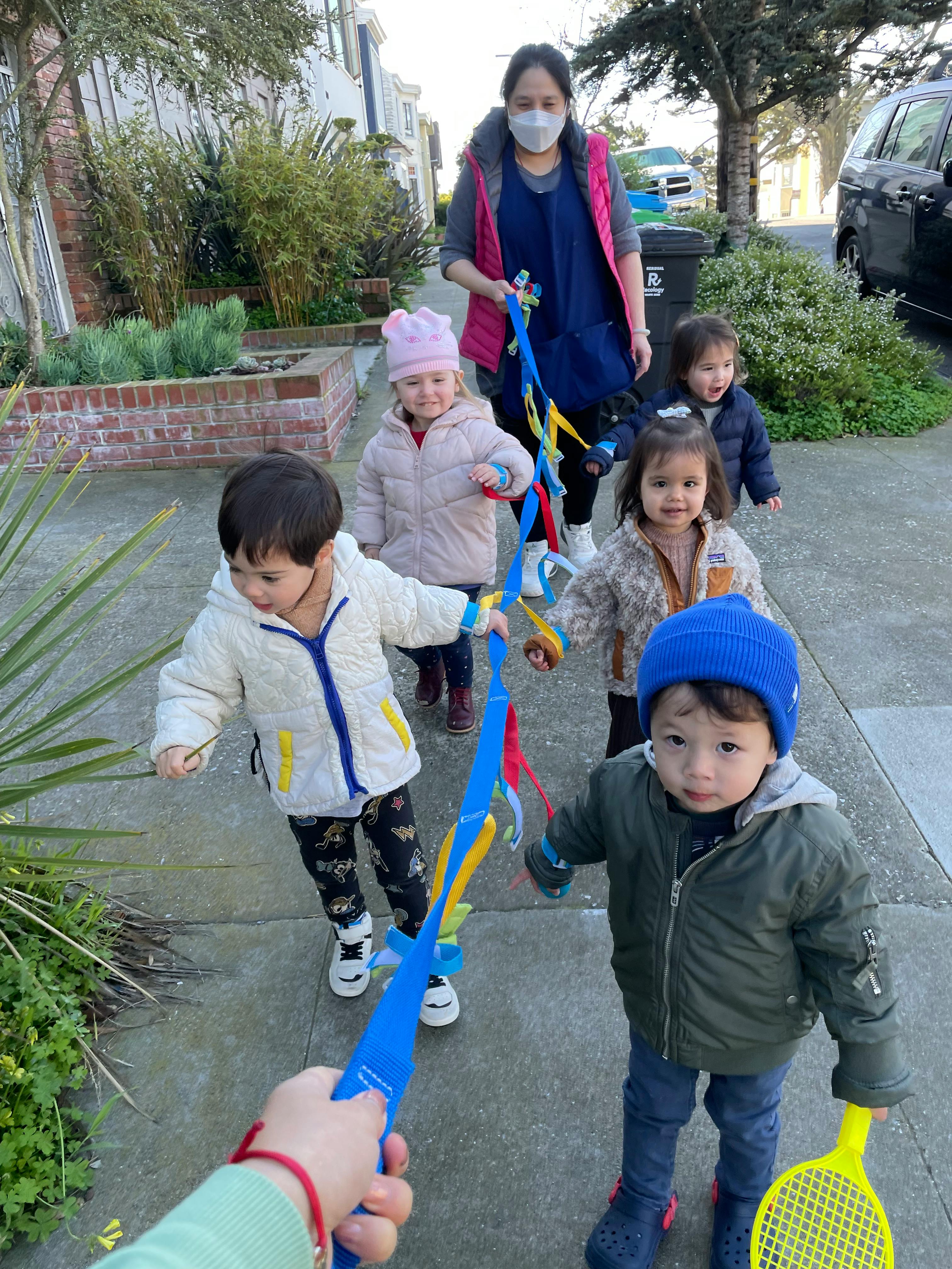 Person in mask walking with four children holding a colorful ribbon