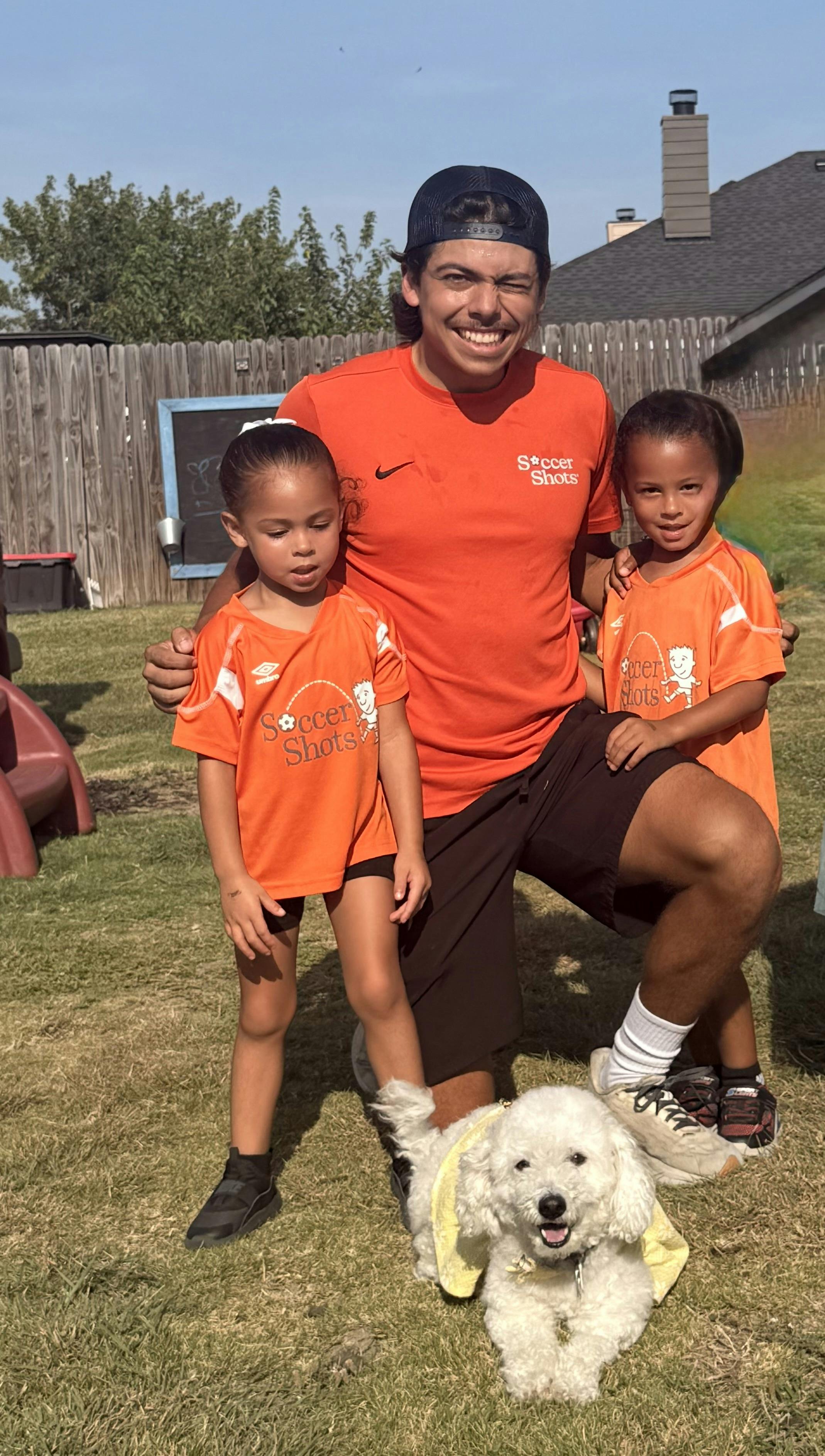 A person and two children in soccer shirts posing outdoors with a small dog
