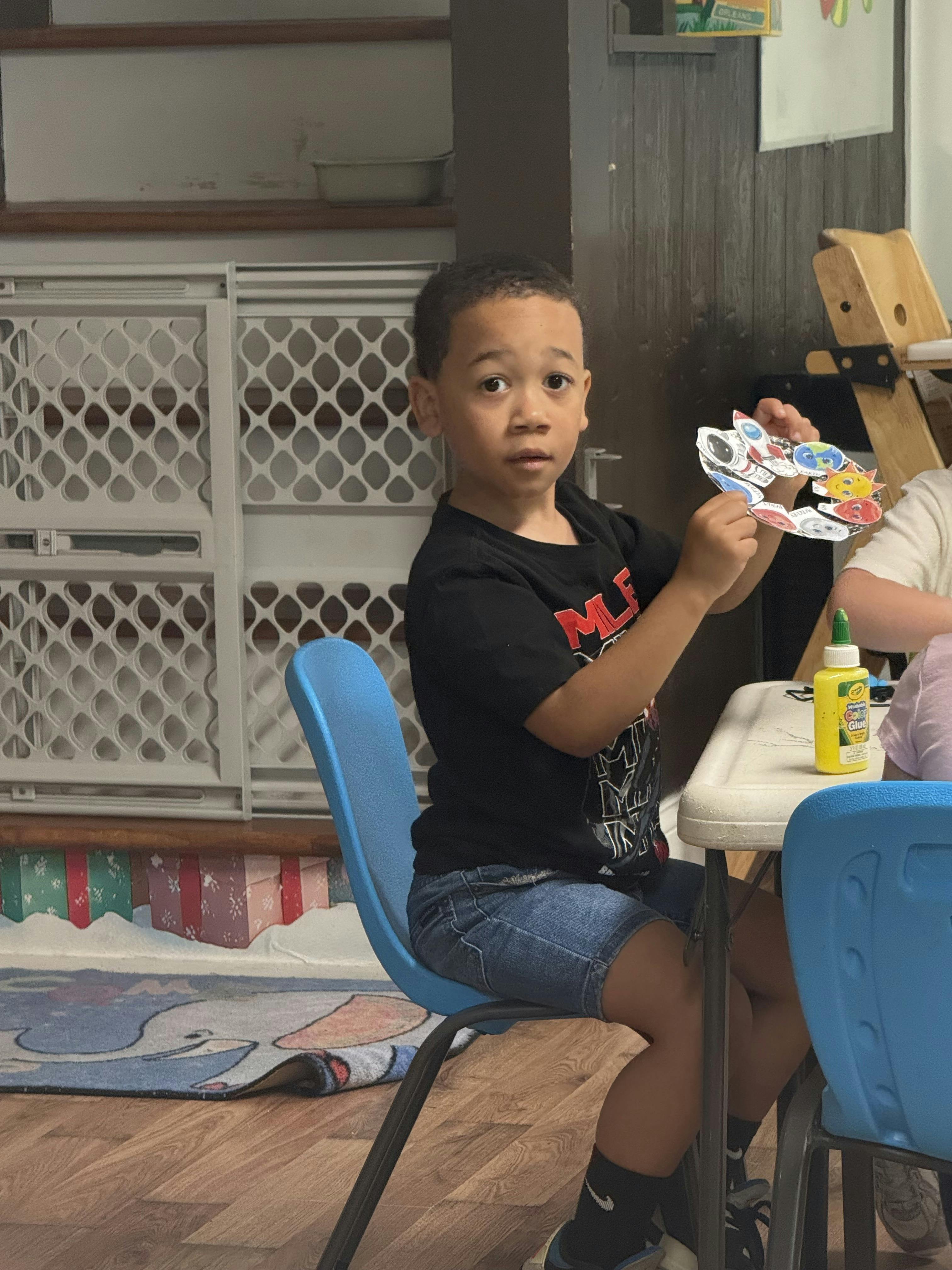 Child seated at a table holding a paper solar system craft