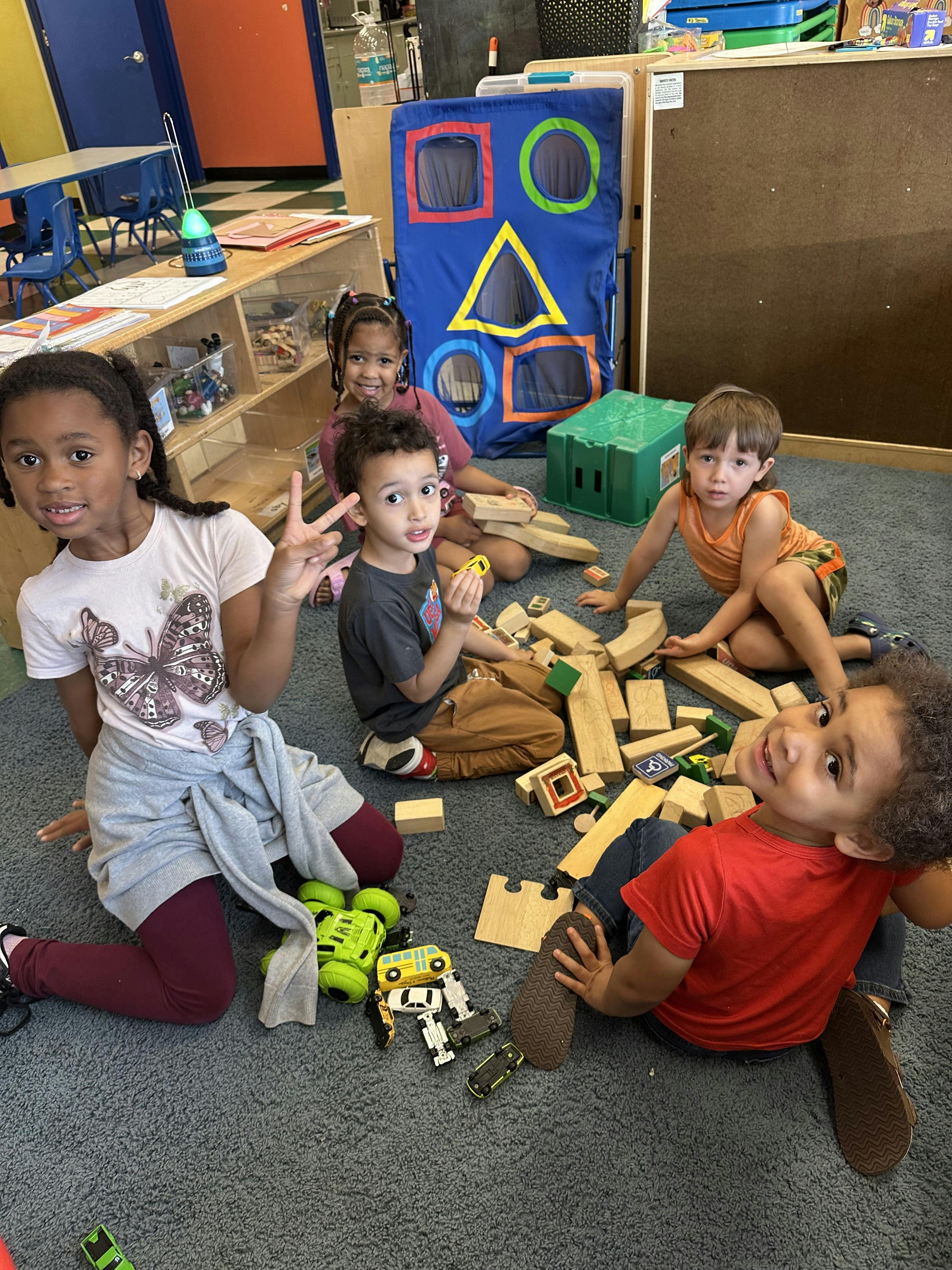 Five children playing with wooden blocks and toy cars on a classroom carpet