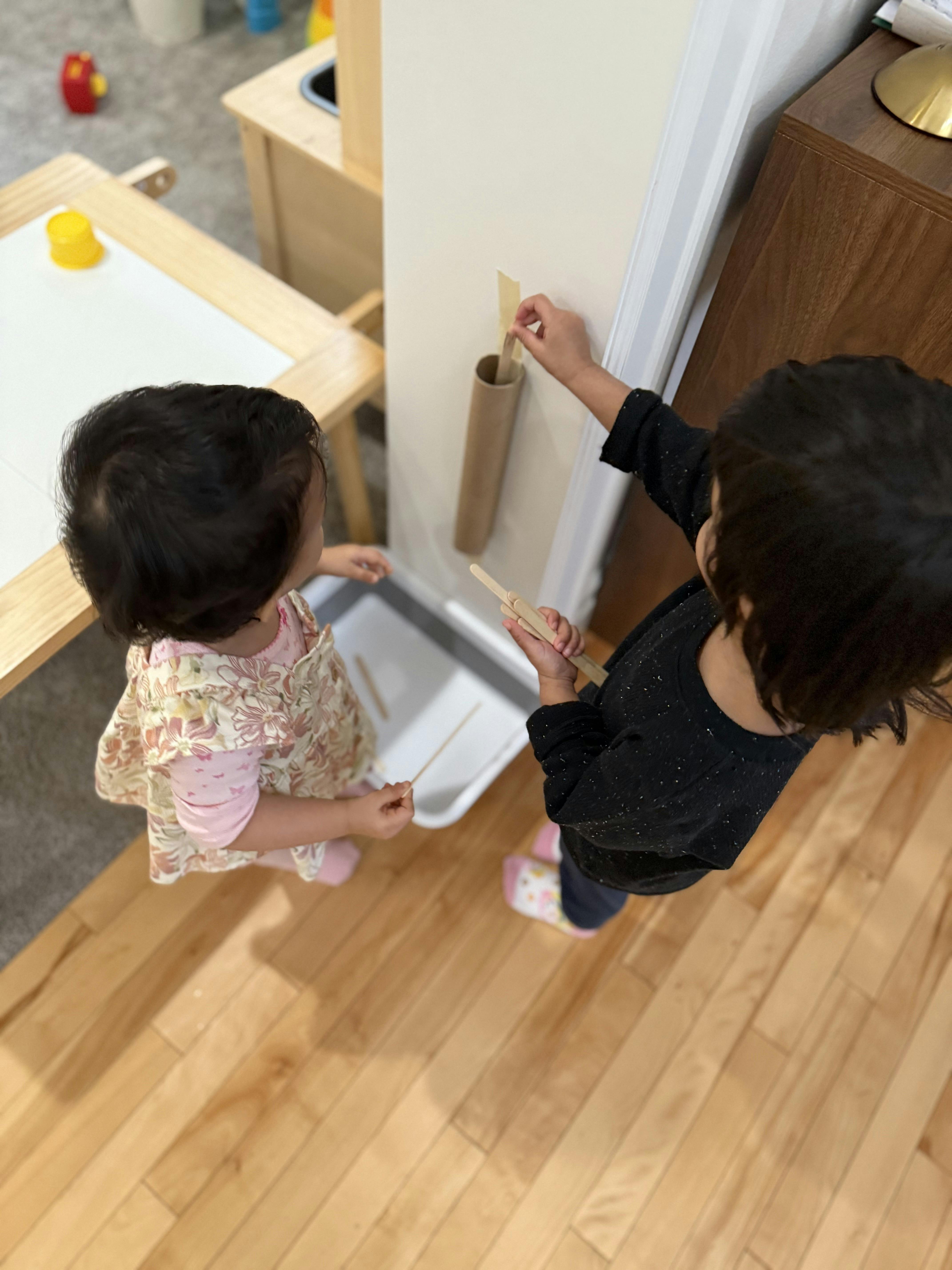 Two young children playing with a cardboard tube taped to a wall and craft sticks