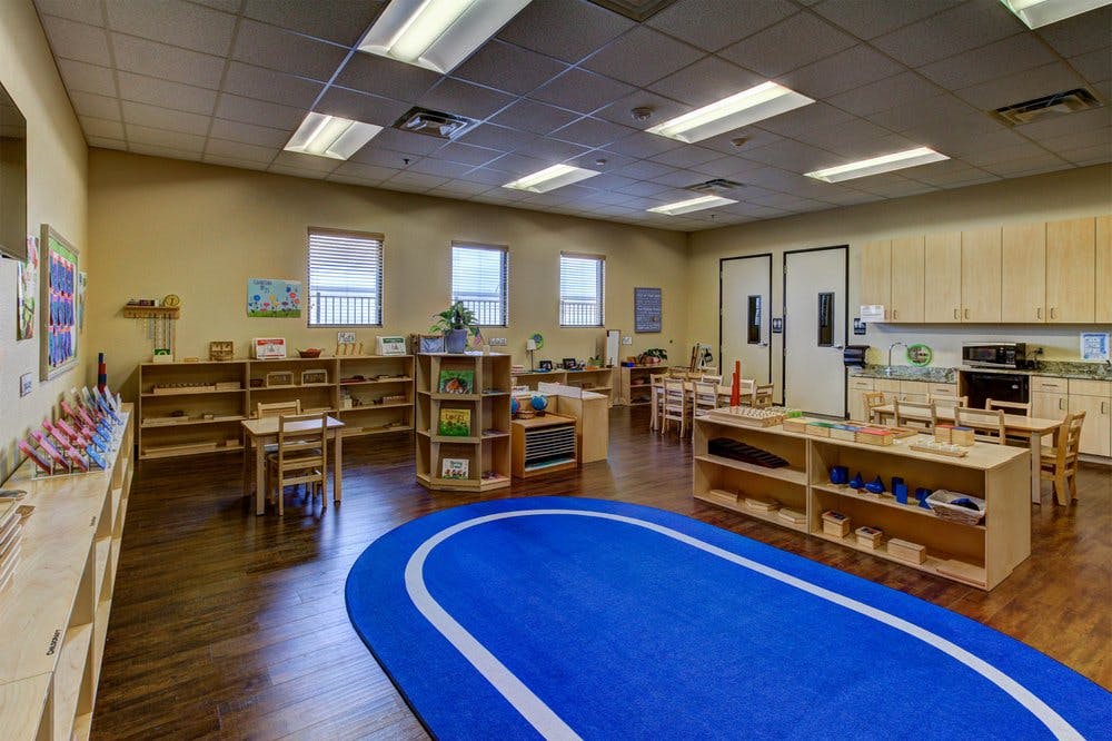 Classroom with small tables and chairs, shelves with learning materials, and a blue rug