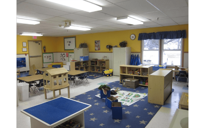 Empty classroom with yellow walls, small tables and chairs, and shelving with toys