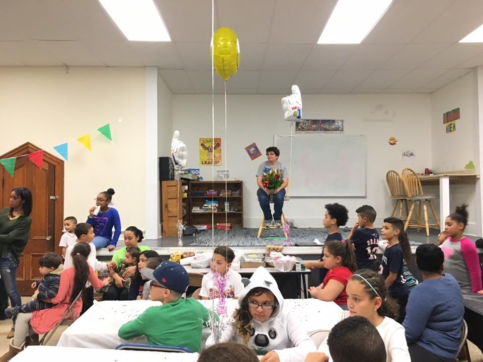 People and children gathered in a room, one person holding flowers on a stage