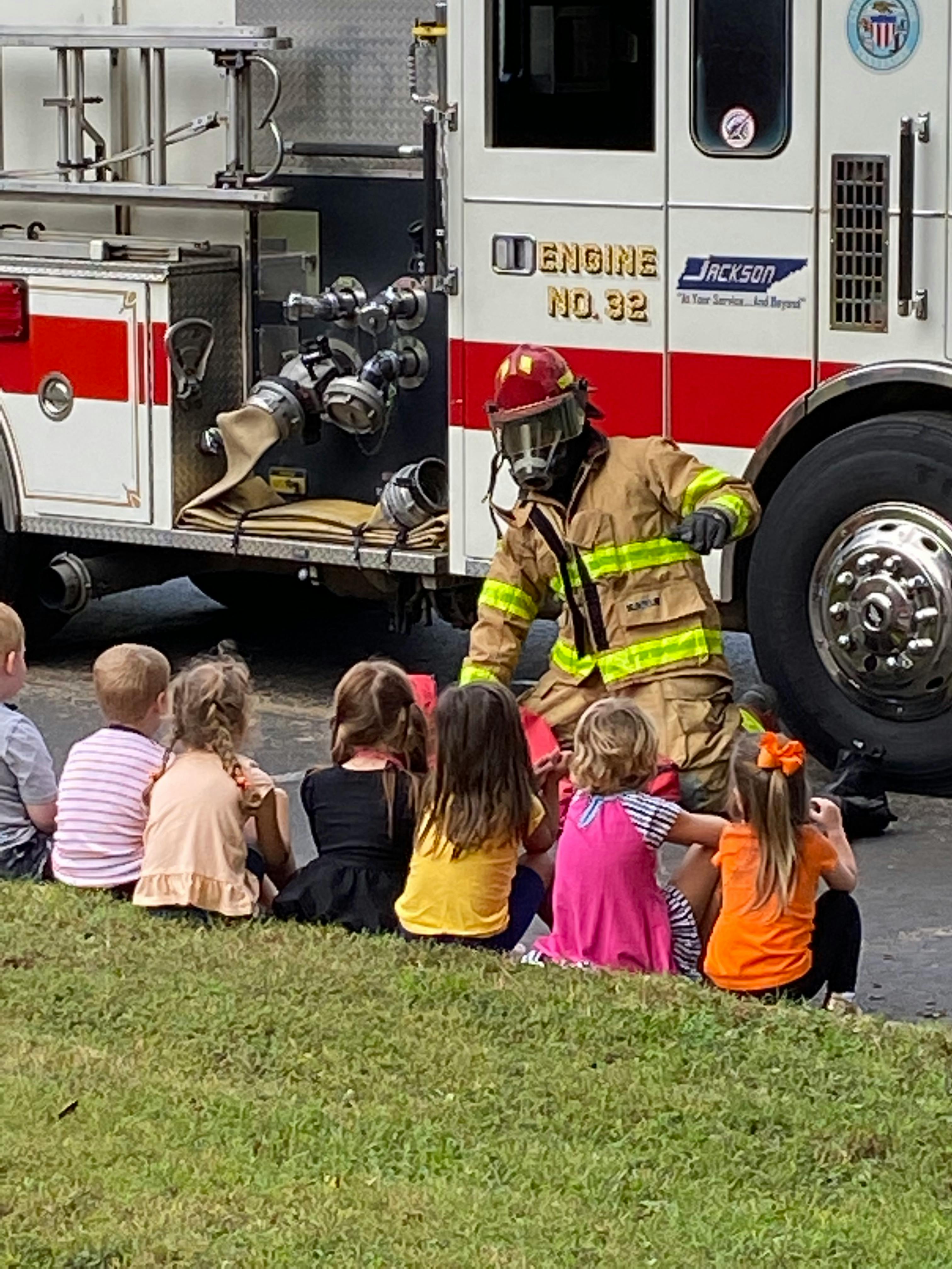 Firefighter in gear kneeling in front of children sitting on grass near a fire truck