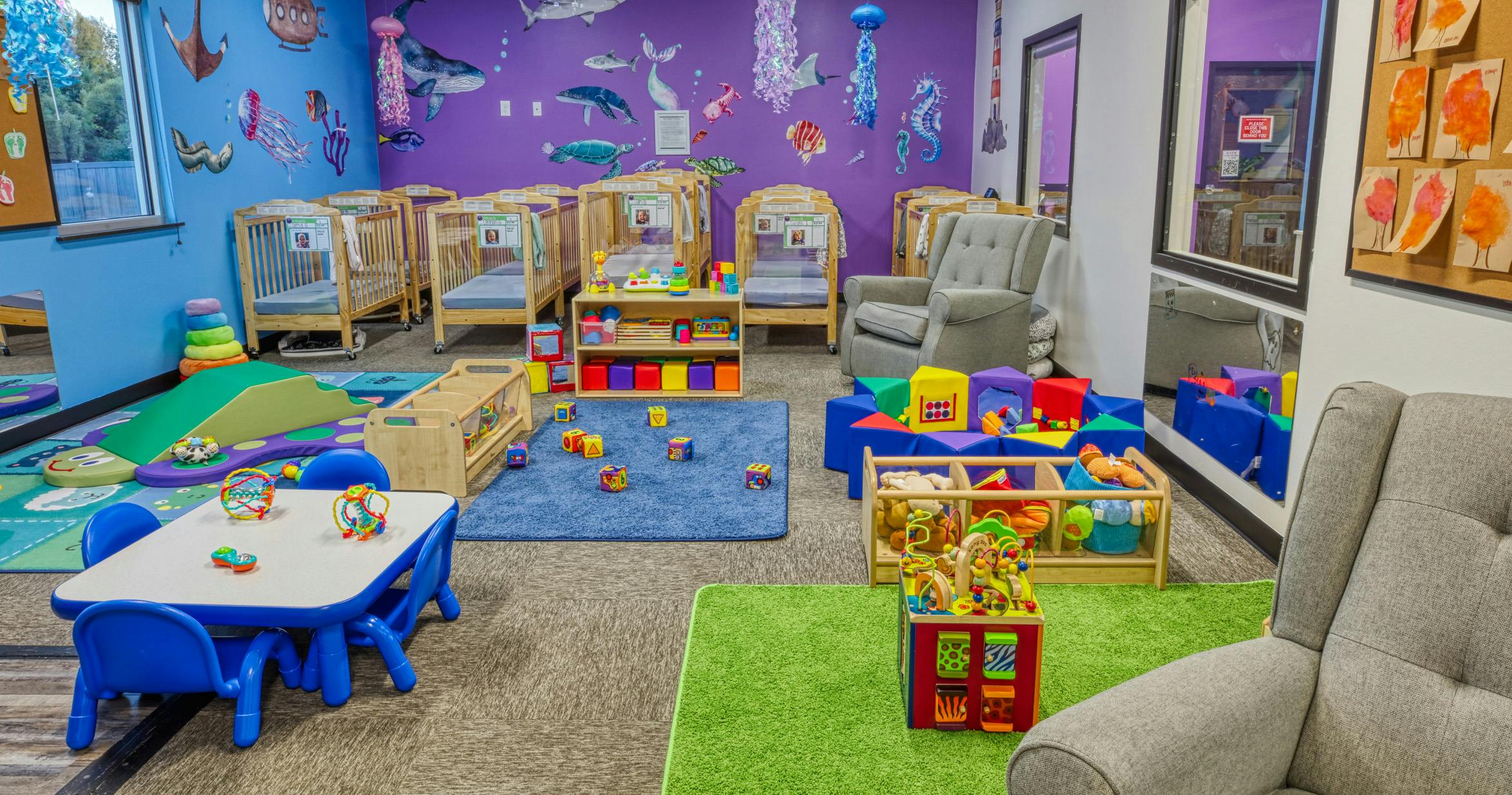 A colorful childcare room with cribs, play mats, toys, and small tables