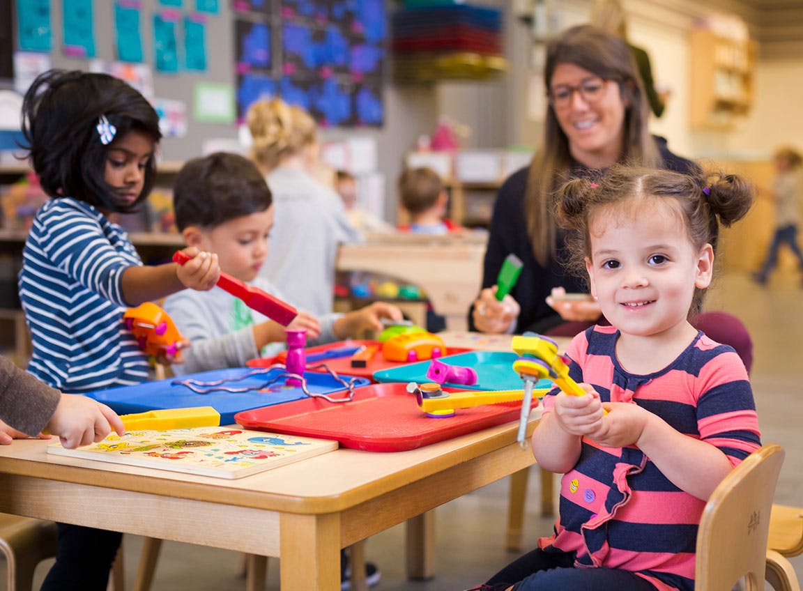 Children playing with toys at a table with an adult in the background