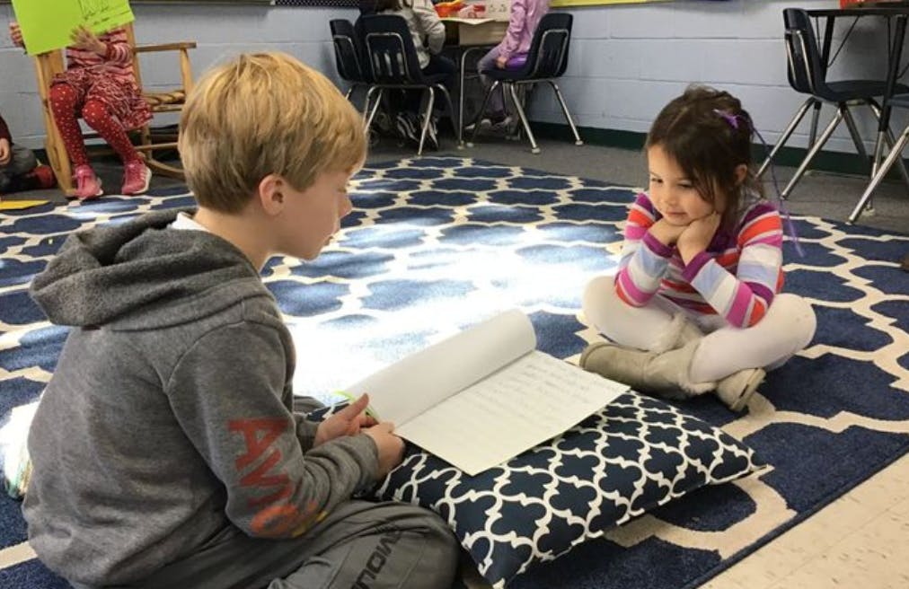 Child reading a book to another child in a classroom