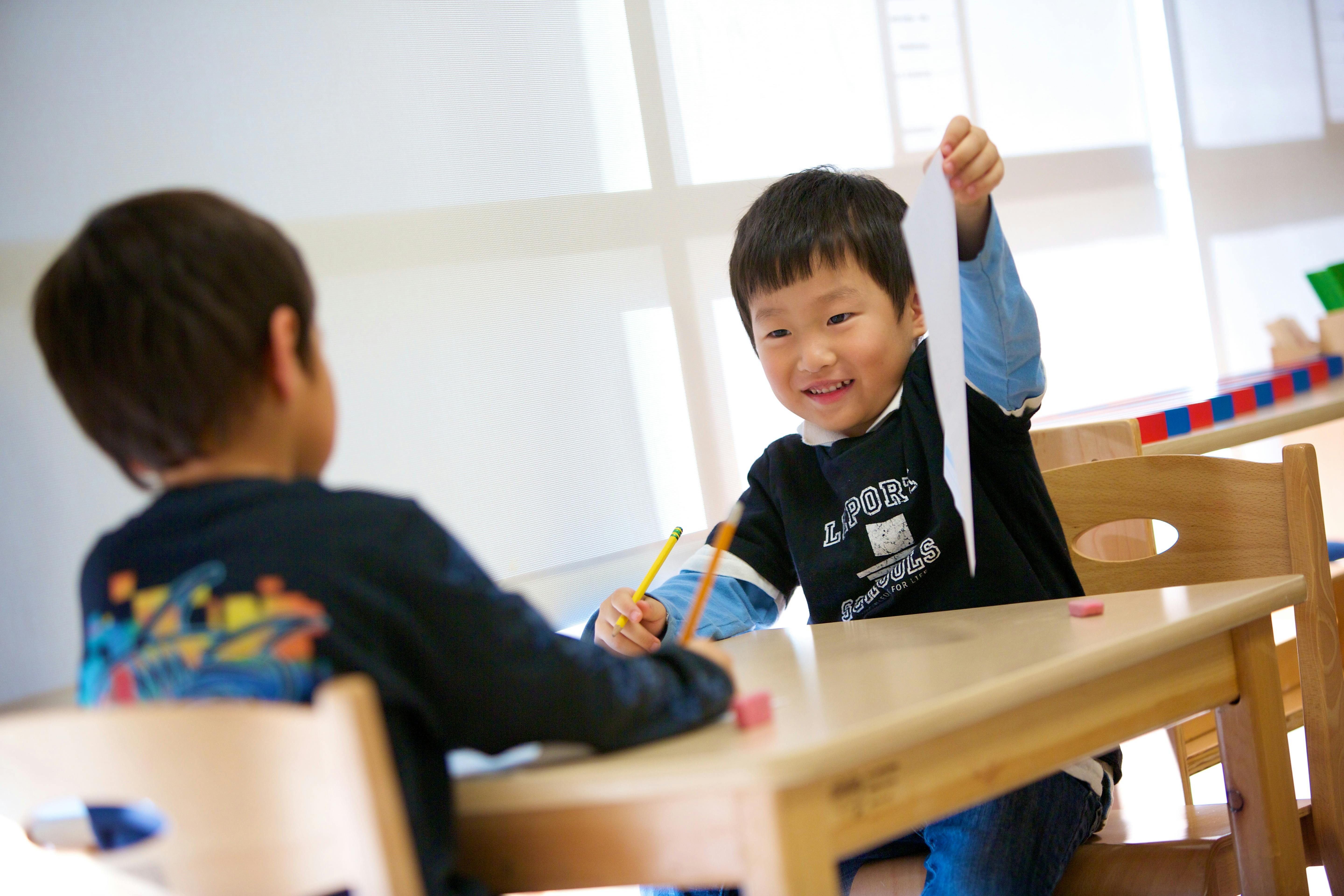Child holding up a piece of paper while sitting at a table with another child
