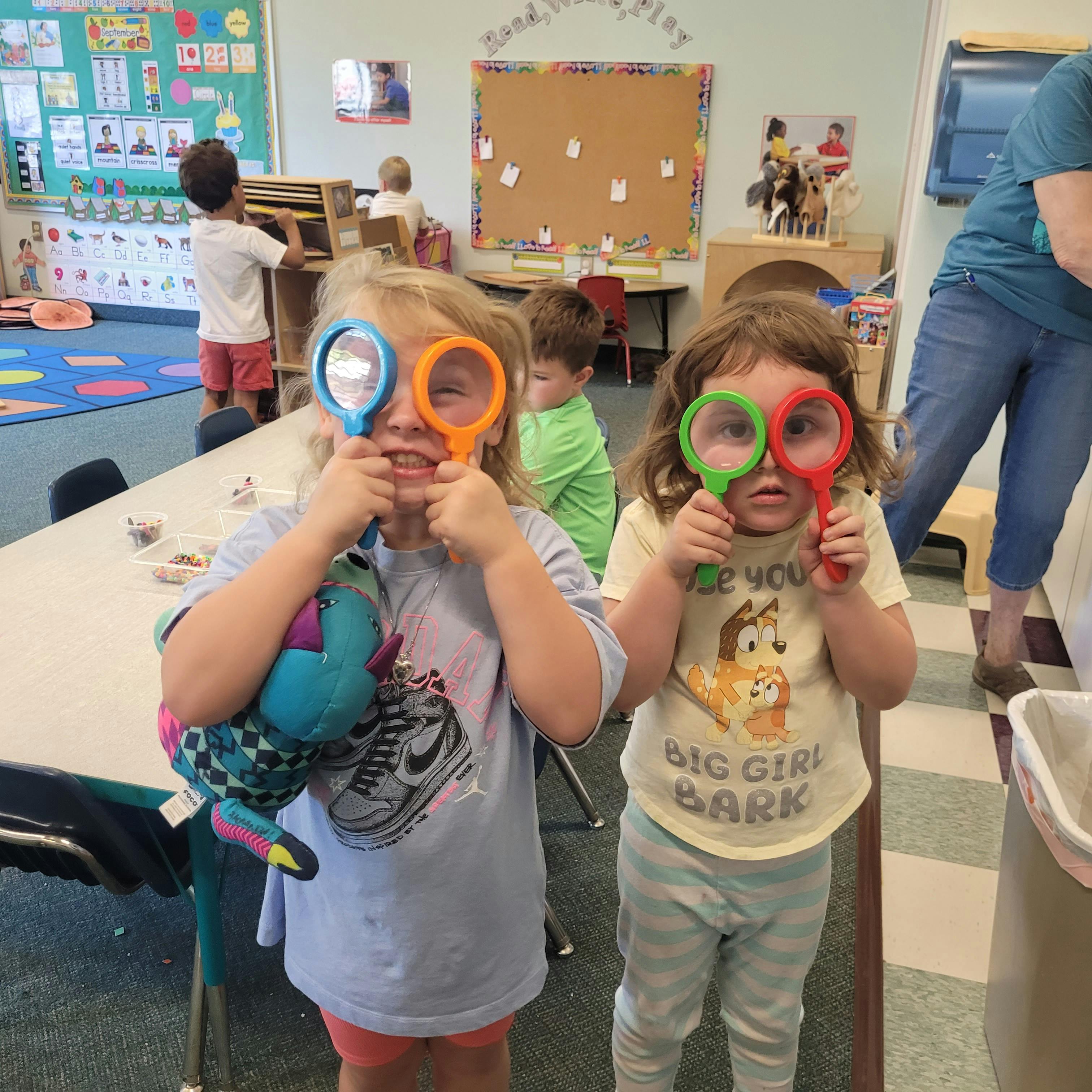 Two children holding magnifying glasses over their eyes in a classroom