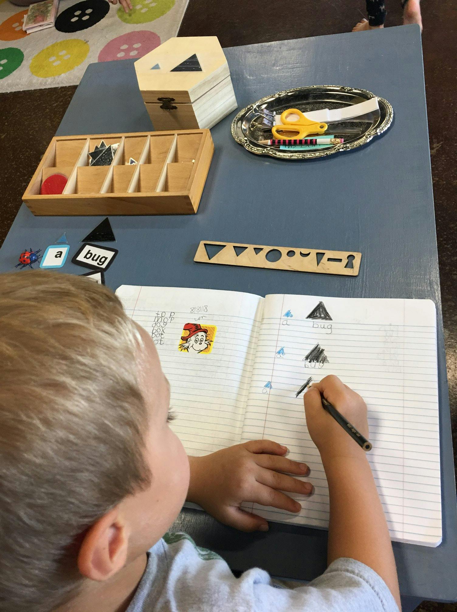 Child drawing shapes in a notebook at a table with learning materials