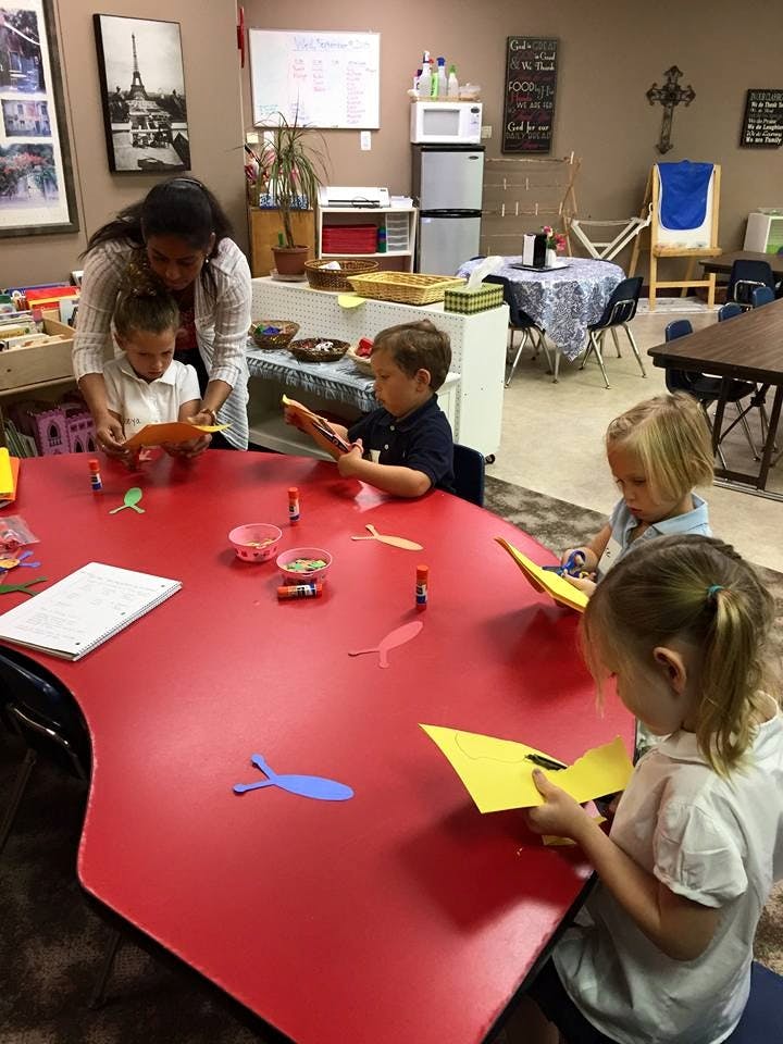 Adult helping children with crafts at a red table in a classroom