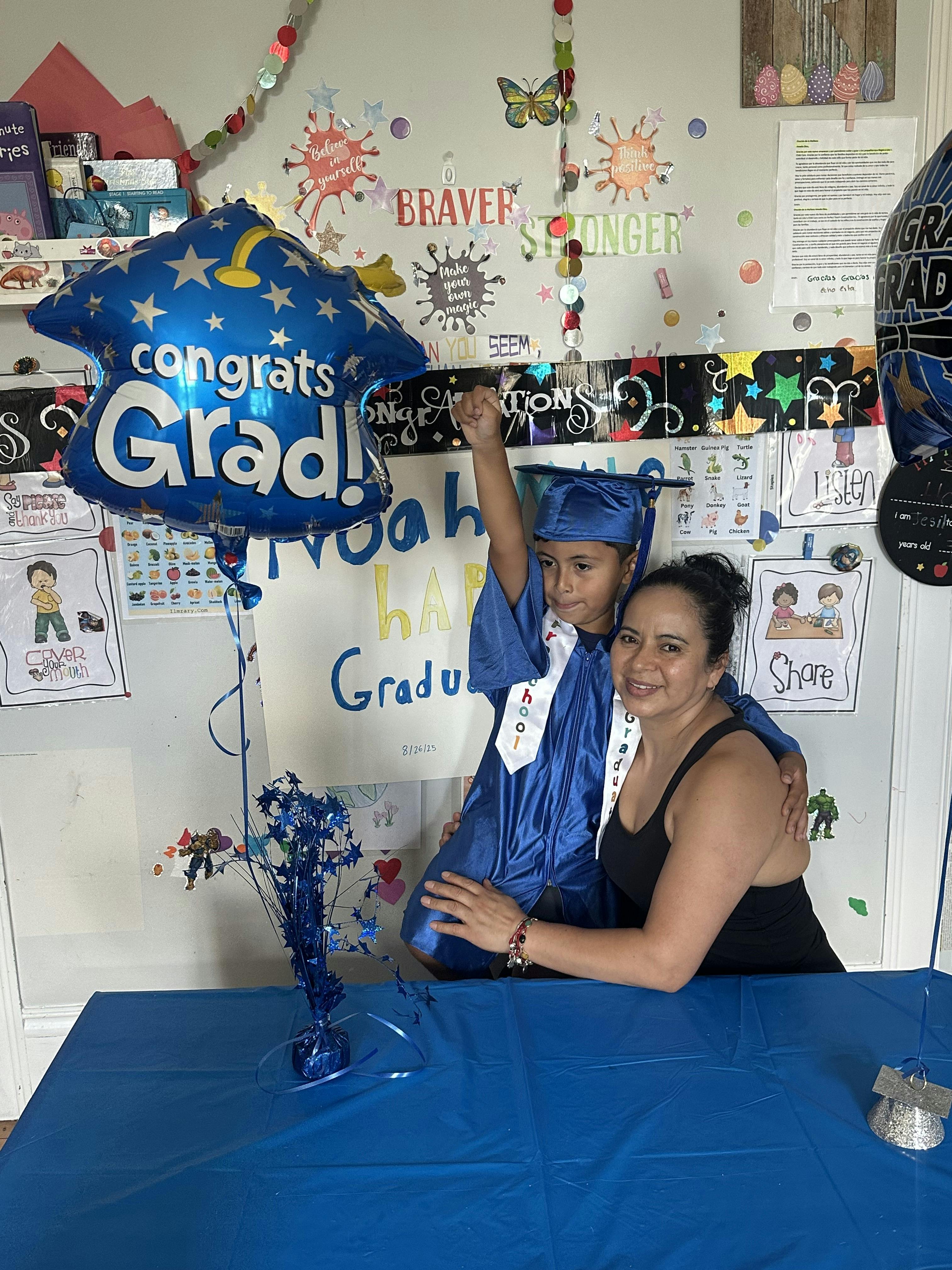 Child in graduation cap and gown with a person, "Congrats Grad" balloon