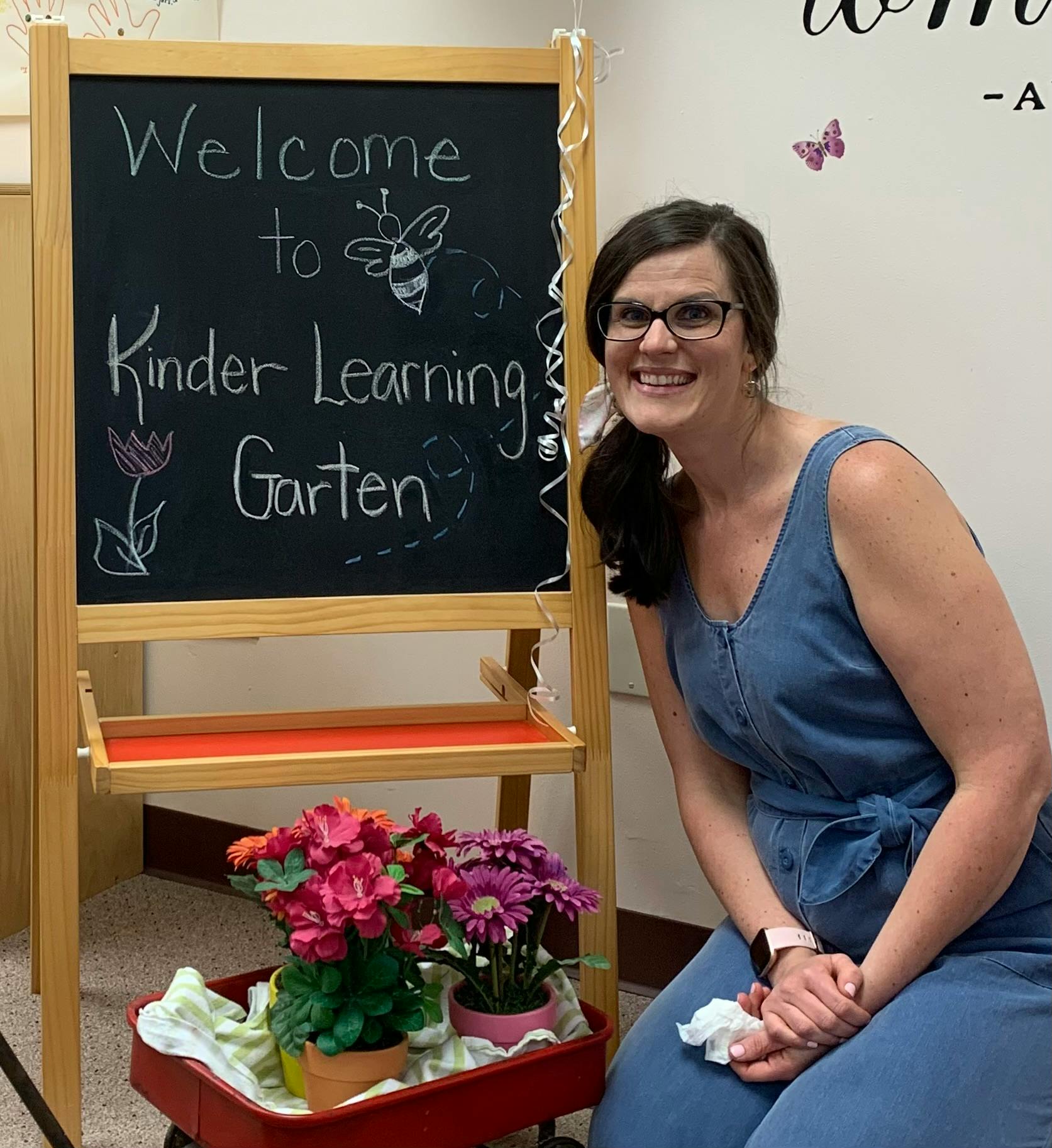 Person kneeling next to a chalkboard easel that says "Welcome to Kinder Learning Garten"